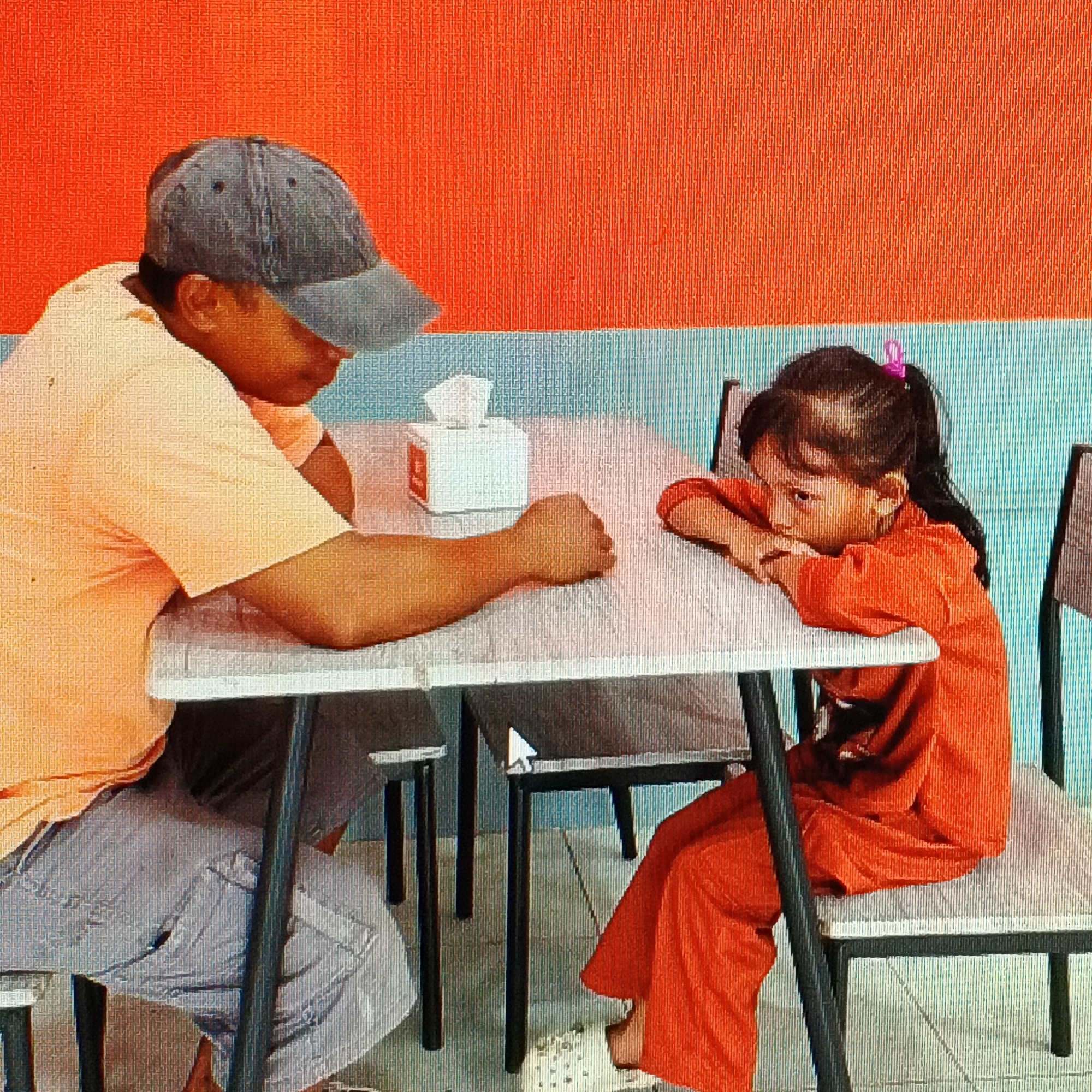 This image depicts an indoor scene, likely a restaurant, with two individuals seated at a table. A person wearing a light orange t-shirt and a grey baseball cap is on the left, with their arm resting on the table. Opposite them, a young girl in an orange outfit is leaning on the table, with her arms crossed. A white tissue box is placed on the table between them. The background consists of a textured orange wall above a light blue striped area. The location is identified as Ende, Indonesia, suggesting this is a captured moment within that city.