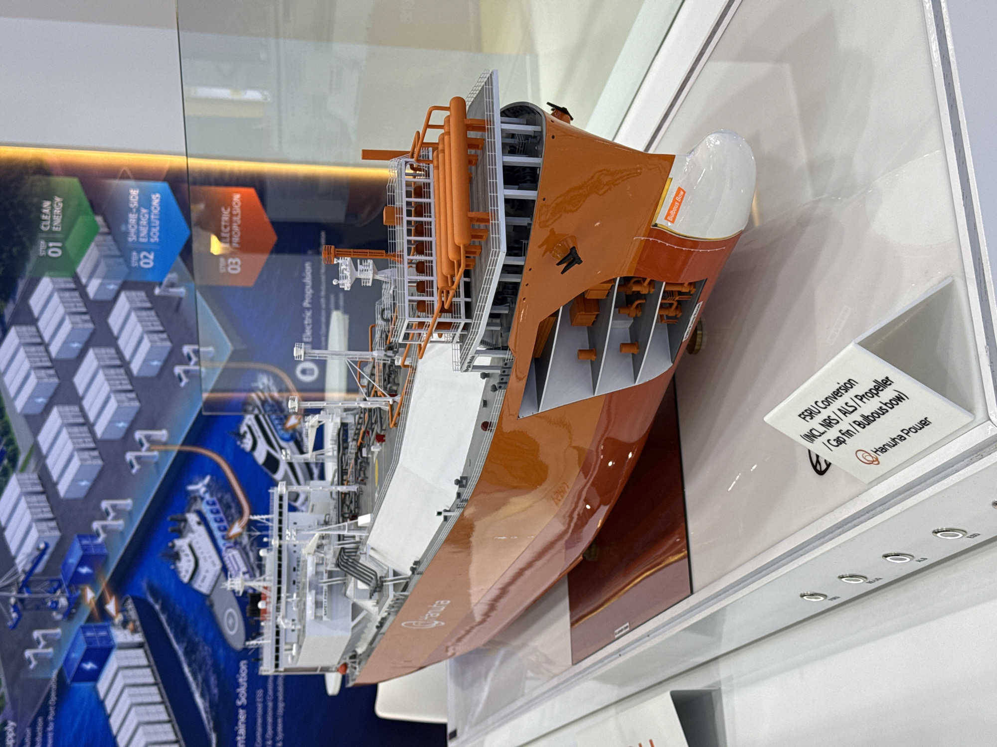 This is a medium shot, eye-level photograph of a scale model of an orange and white ship, likely a liquefied natural gas (LNG) carrier, displayed at an exhibition. The model is positioned diagonally in the frame, with its bow pointing towards the right and slightly towards the viewer. The ship model is detailed, showing internal compartments with machinery and piping, as well as the deck with various structures. The name "Hanwha" is visible on the hull.
Behind the ship model, a large graphic display illustrates a system of clean energy solutions, with three steps labeled: "01 CLEAN ENERGY," "02 SHORE-SIDE ENERGY SOLUTIONS," and "03 ELECTRIC PROPULSION." The graphic shows container ships, cranes, and smaller vessels.
In the foreground, a white display stand with the text "FSRU Conversion (INCL. NRS / ALS / Propeller / Cap fin / Bulbous bow) Hanwha Power" is visible. The model rests on a white platform, which is part of a larger display. The background is out of focus, showing glass or a reflective surface, and what appears to be other exhibition booths.
The lighting is bright, suggesting an indoor exhibition environment. The photograph is taken in Bangkok, Thailand.