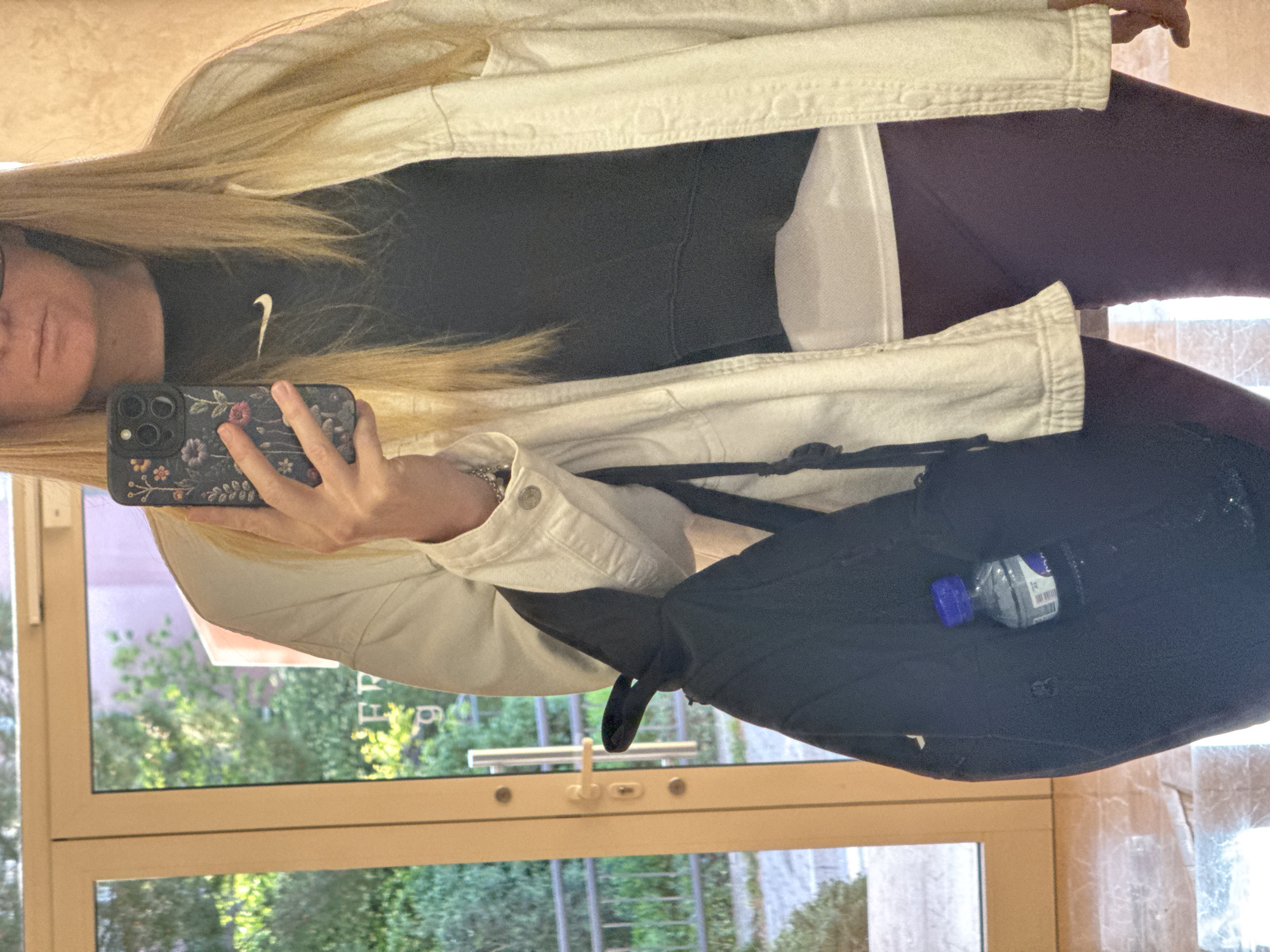 The image captures a person taking a mirror selfie indoors in Budva, Montenegro, under bright daytime conditions. The subject, who has long blonde hair, is wearing a casual outfit consisting of a light-colored, possibly off-white, denim jacket over a black top. The white "Nike" swoosh logo is clearly visible on the chest of the black top. They are also wearing dark, possibly maroon or purple, leggings.
The person is holding a smartphone with a distinctive dark floral-patterned case in their right hand, angled towards the reflective surface. Only the lower portion of their face, including the mouth and chin, is visible, partially obscured by the phone. A black backpack is slung over their shoulder, and a water bottle with a blue cap is visible inside.
The background reveals a large window looking out onto a vibrant outdoor scene with abundant green trees and bushes, suggesting a pleasant, possibly sunny, day. Faint, reversed text, potentially "EFT" or "FFF", is barely discernible in the window's reflection. The indoor floor or wall, where the selfie is being taken, is highly reflective, creating the mirror effect.