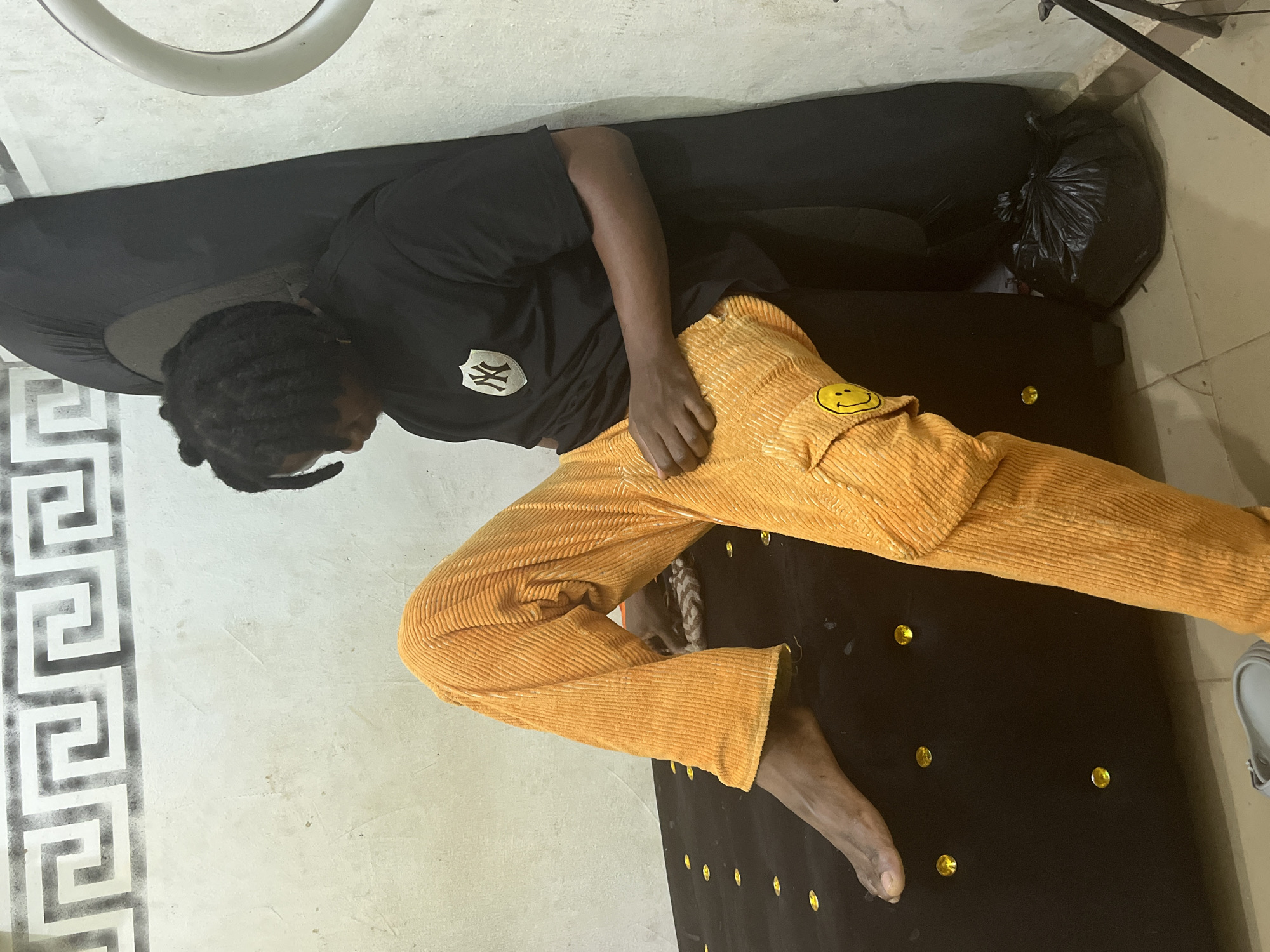 This image depicts an indoor scene in Jos, Nigeria, featuring a young individual seated on a dark, upholstered surface adorned with scattered yellow gem-like studs. The person, with dark skin and dreadlocked hair, wears a black short-sleeved t-shirt featuring a white shield emblem with a stylized 'NY' logo on the chest, and bright orange corduroy pants with a yellow smiley face patch on the right cargo pocket. They are barefoot, with their right foot resting on the upholstered surface. The individual is looking downwards, with their hands resting on their lap. The background consists of a light-colored, possibly off-white, wall displaying a repeating grey Greek key pattern stenciled along the top. Leaning against the wall behind the person is a large, curved, dark object, likely a padded panel or cover. To the far right, a white circular ring light is partially visible, with a portion of a black tripod stand extending from the bottom right corner. A full black plastic trash bag rests on the tiled floor in the lower right foreground. A grey slide sandal is partially visible on the floor in the lower left.