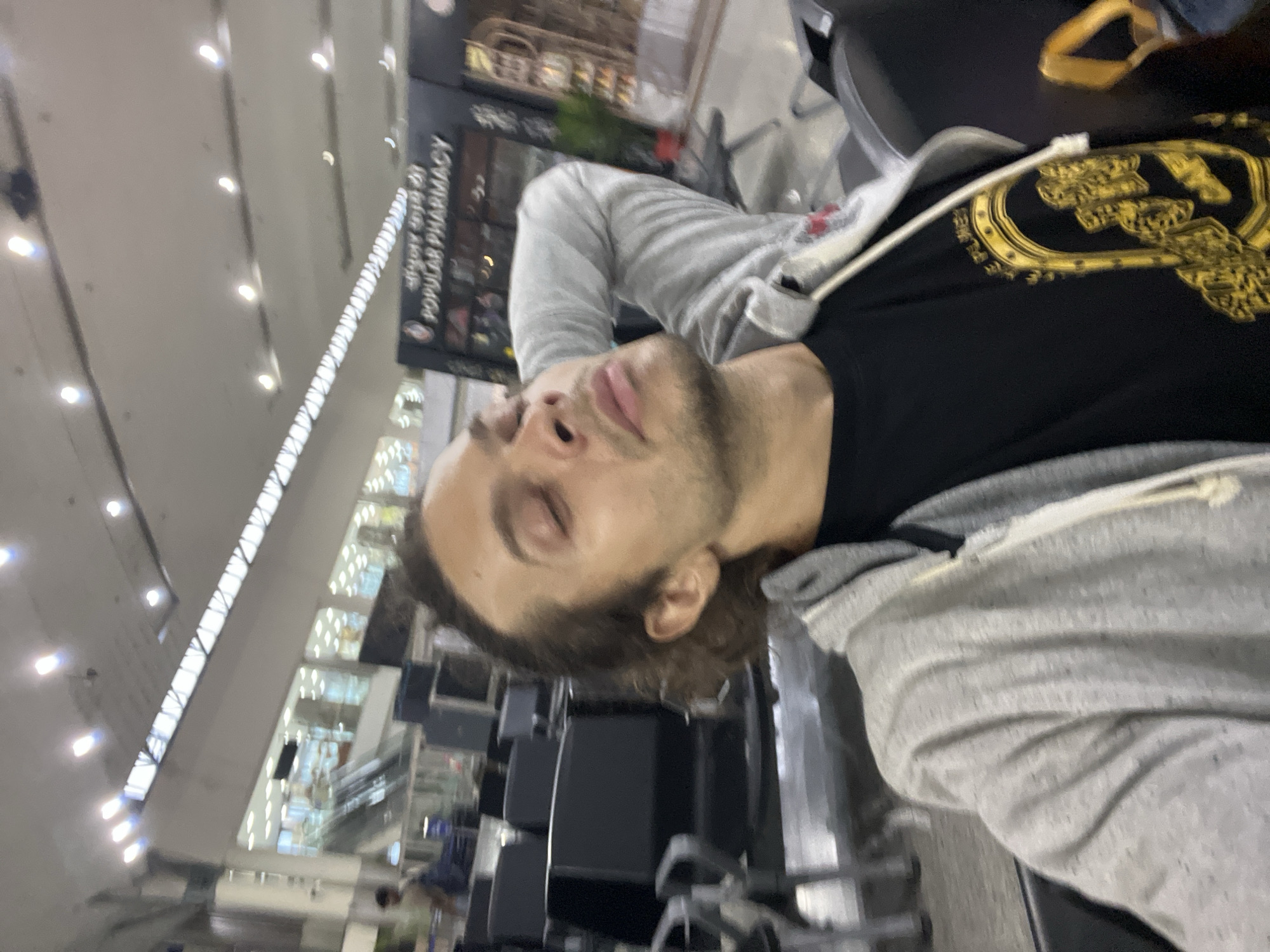 The photograph appears to have been taken in an airport terminal. A man with light skin, brown hair, and stubble lies sideways on chairs. He is wearing a grey hoodie over a black t-shirt with a gold design. The terminal has rows of unoccupied seating, and in the background, there's a sign that says "Popular Pharmacy". The location is likely Lal Bahadur Shastri International Airport, Babatpur, India.