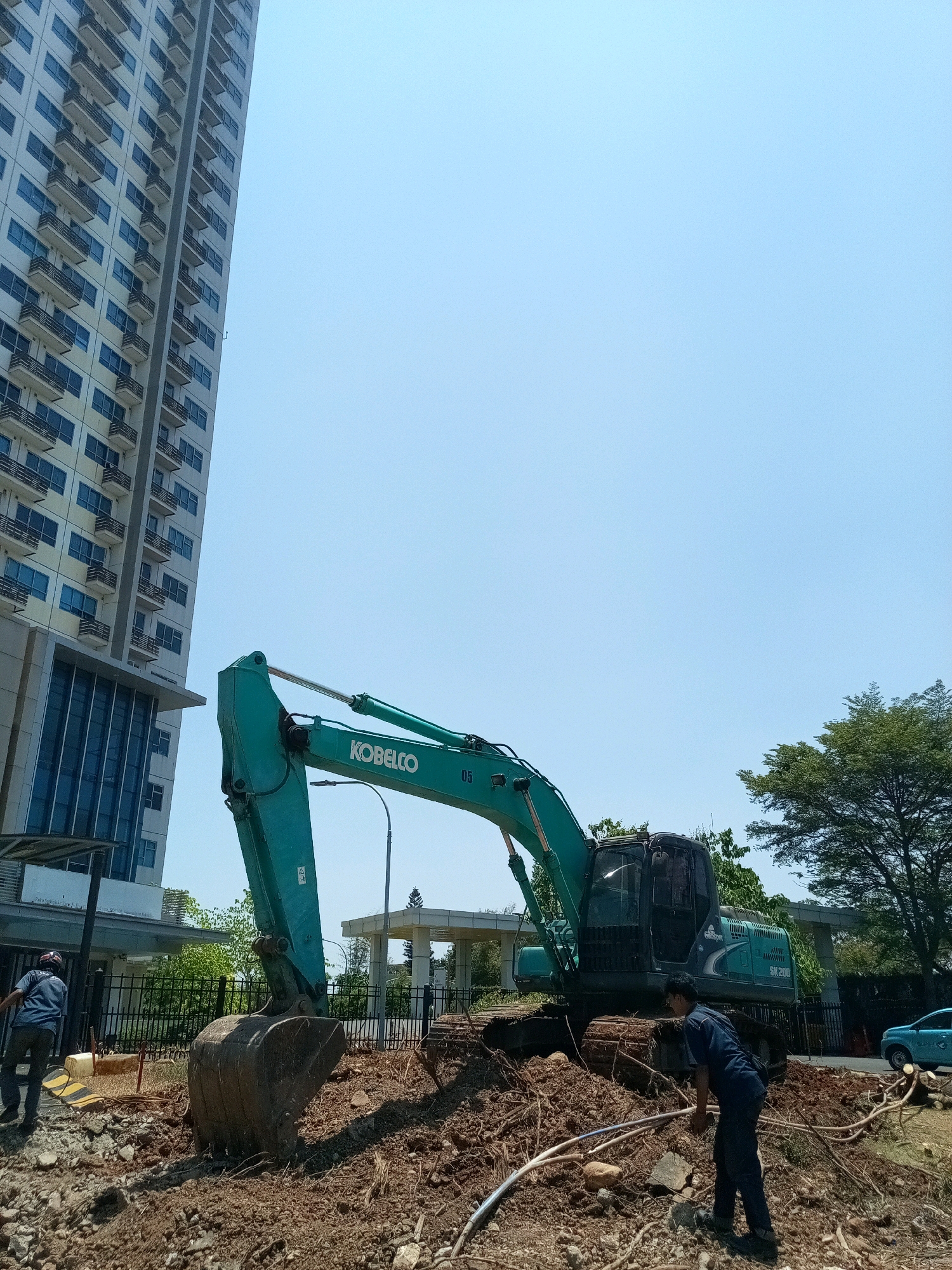 In Bekasi, Indonesia, a construction site is depicted under a clear blue sky. A tall, modern apartment building with numerous balconies and blue-tinted windows dominates the left side of the frame. In the foreground, a large green Kobelco excavator with the model number SK200 is positioned on a mound of dirt and debris. Its large bucket is filled with soil. Two individuals are present; one, wearing a blue shirt and dark pants, is operating the excavator, while another, in a blue uniform, is standing near the excavator, appearing to be engaged in some work with the debris. The background shows a street with a light-colored car and some lush green trees, suggesting an urban or suburban setting.