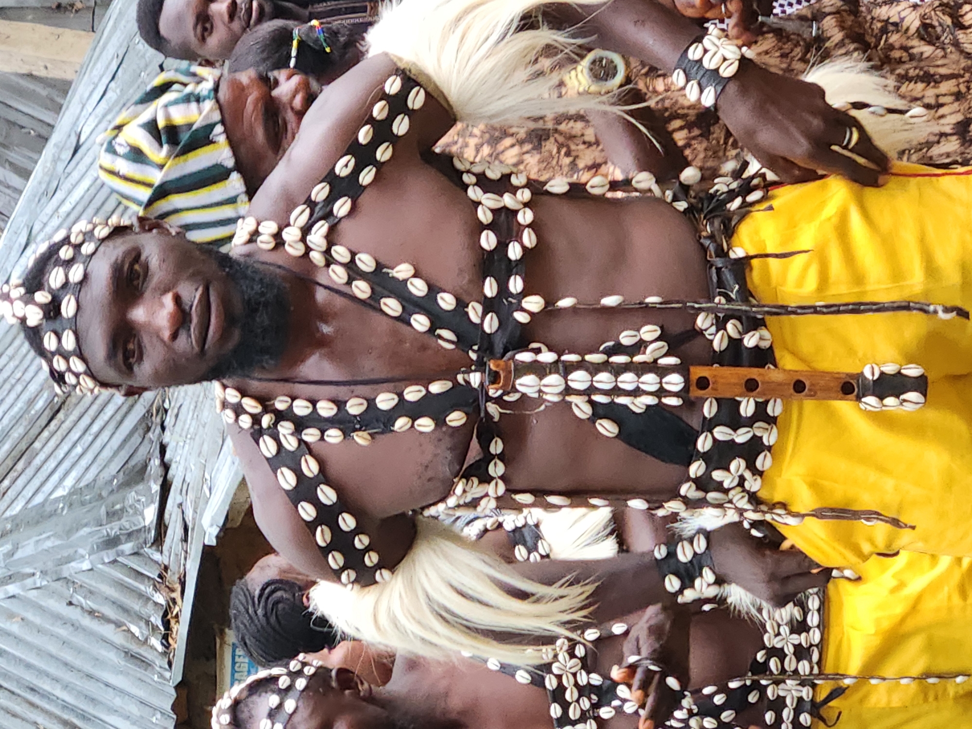 In an outdoor setting, a group of people are gathered, with a focus on a man adorned in traditional attire. He wears a headdress and chest ornamentation made of seashells and black material, with a wooden flute hanging from his chest. His attire also includes a yellow garment and fluffy white accents on his arms. Another person to his left wears a similar seashell-adorned headdress, while a man to his right wears a striped cap and is partially obscured. The background shows a corrugated metal roof structure and glimpses of other individuals, suggesting a cultural event or ceremony.