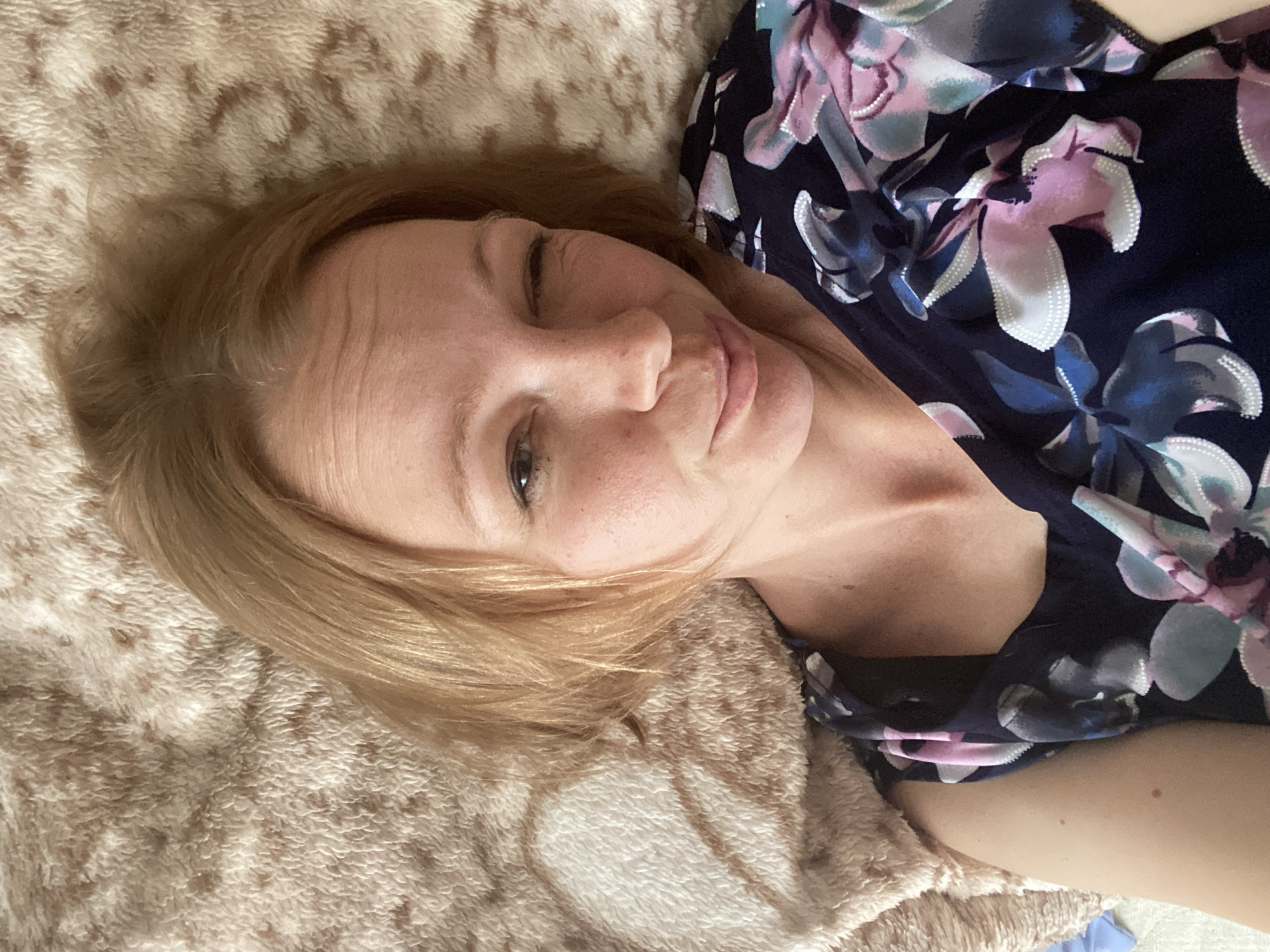 An overhead, close-up shot shows a woman lying on a beige, fuzzy blanket and winking at the camera. The woman has fair skin, auburn hair, and is wearing a navy blue dress with a floral pattern of pink, blue, and white flowers. The blanket has a pattern of what appear to be bear outlines. The scene appears to be indoors. The city of Maykop, Russia is not depicted in this image.