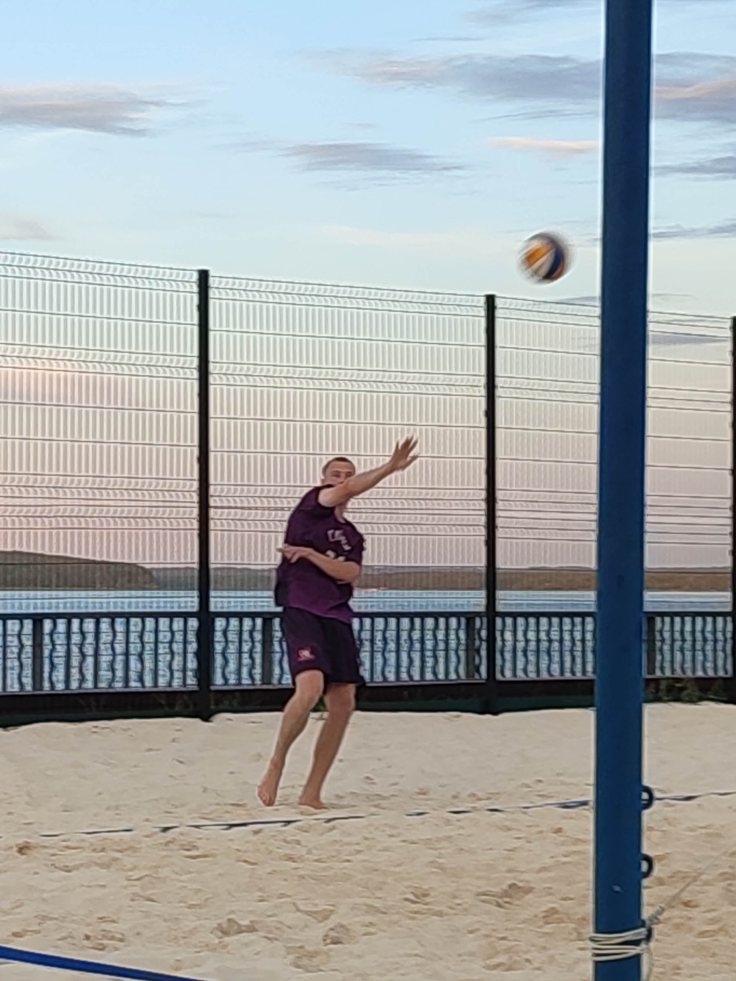An unidentified male is playing beach volleyball. He is wearing a purple t-shirt and dark shorts. He is in the middle of an action, possibly about to hit the ball, which is in the air above him. The game is being played on a sand court, with a net in the foreground. In the background, there is a fence, a body of water, and a cloudy sky. The overall lighting suggests it is either early morning or late evening.
