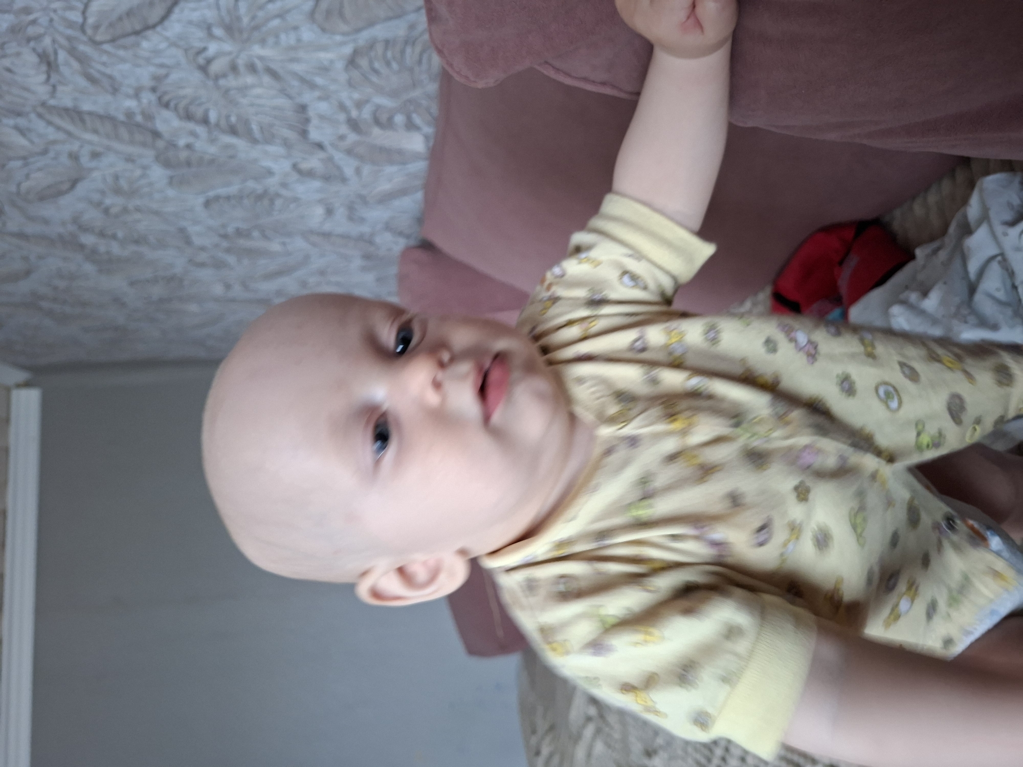 The image features a close-up, upside-down shot of a bald baby lying on its back. The baby is wearing a yellow pajama set adorned with cartoon animal prints. Its left arm is raised and resting on a pink upholstered surface, likely a sofa or chair. The baby's face is turned towards the camera, with its eyes open and looking slightly to the left. Its mouth is open, as if speaking or about to speak. The background is blurred, but hints of a patterned wallpaper and a wall can be seen. The overall lighting suggests an indoor setting, possibly during daylight hours. There is no visible text in the image. The provided location context is Karsha, Russia.