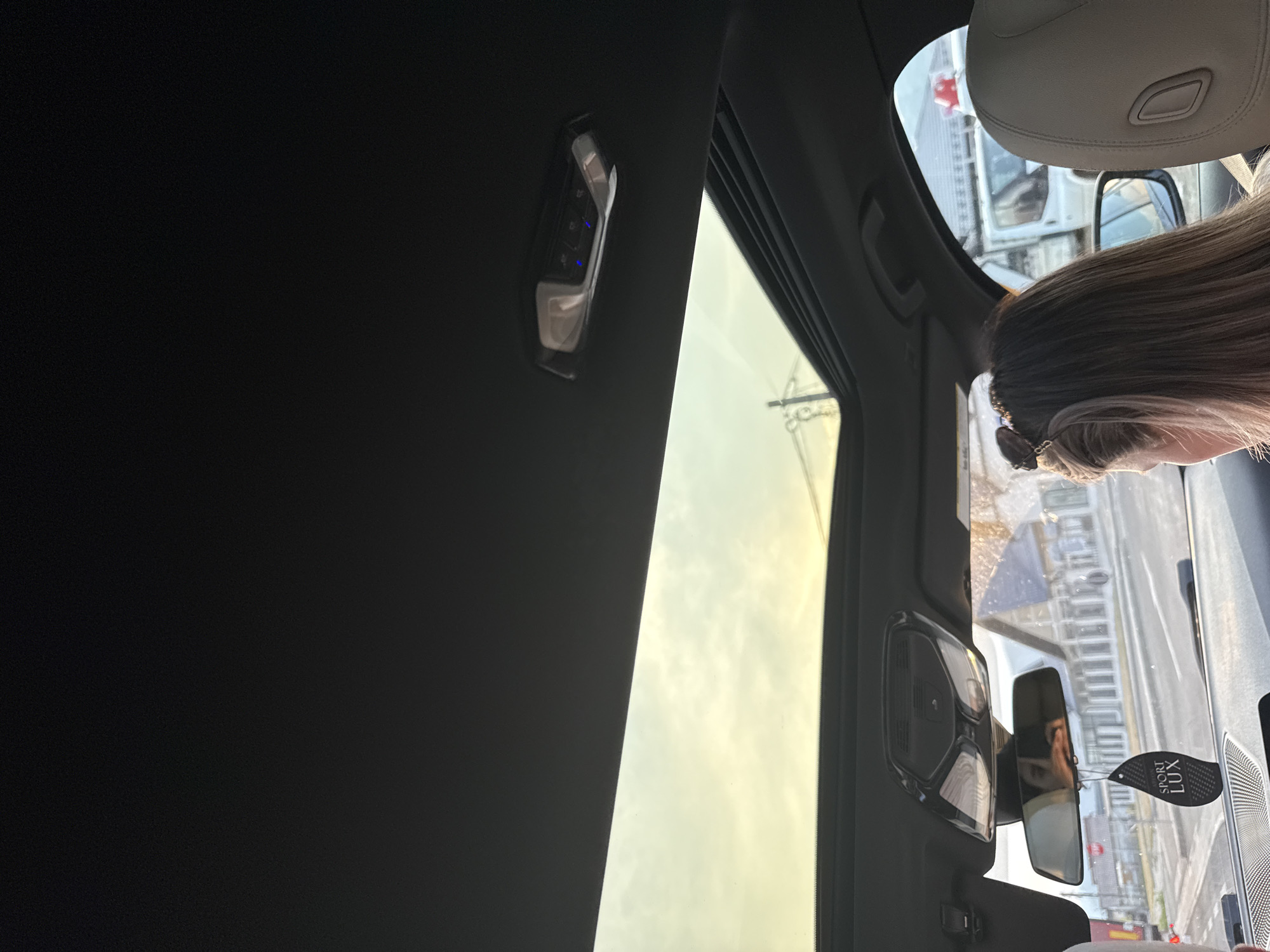 The image, taken from inside a vehicle, captures a woman with long, blonde hair sitting in the passenger seat, facing forward and slightly to the right. She is wearing a light-colored top. Her hair is partially pulled back with sunglasses perched on top of her head. In the foreground, a black car air freshener with the words "SPORT LUX" is hanging from the rearview mirror.
The vehicle has a sunroof, which is open, revealing a pale yellow sky with clouds. The interior of the car is visible, including part of the dashboard and the headliner with interior lights. The rearview mirror reflects the woman's face and the scene outside the car, showing buildings and a road.
Outside the car, a road is visible with some buildings and a white van. The context of Oituz, Romania, is a crucial detail provided for this scene. The image depicts a moment during what appears to be a drive.