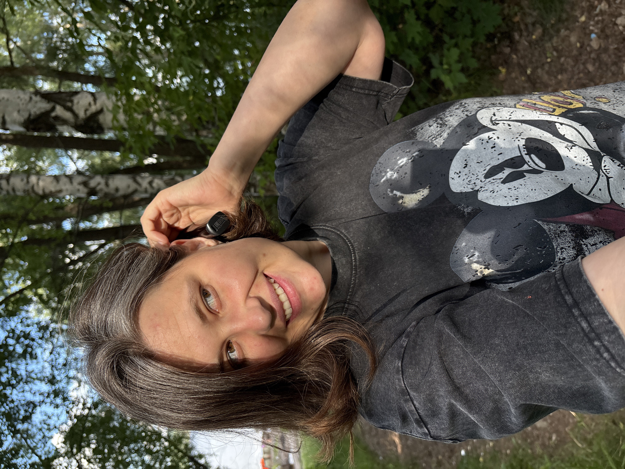 A woman is pictured from the chest up, lying on her back and looking at the camera. She is outdoors, with trees and foliage in the background. The lighting suggests it is daytime, with dappled sunlight filtering through the leaves. She has shoulder-length brown hair and is wearing a dark grey t-shirt with a faded Mickey Mouse graphic on it. She is holding her right ear with her left hand, and appears to be smiling. The overall mood is relaxed and casual. The image was taken in Chaikovsky, Russia.