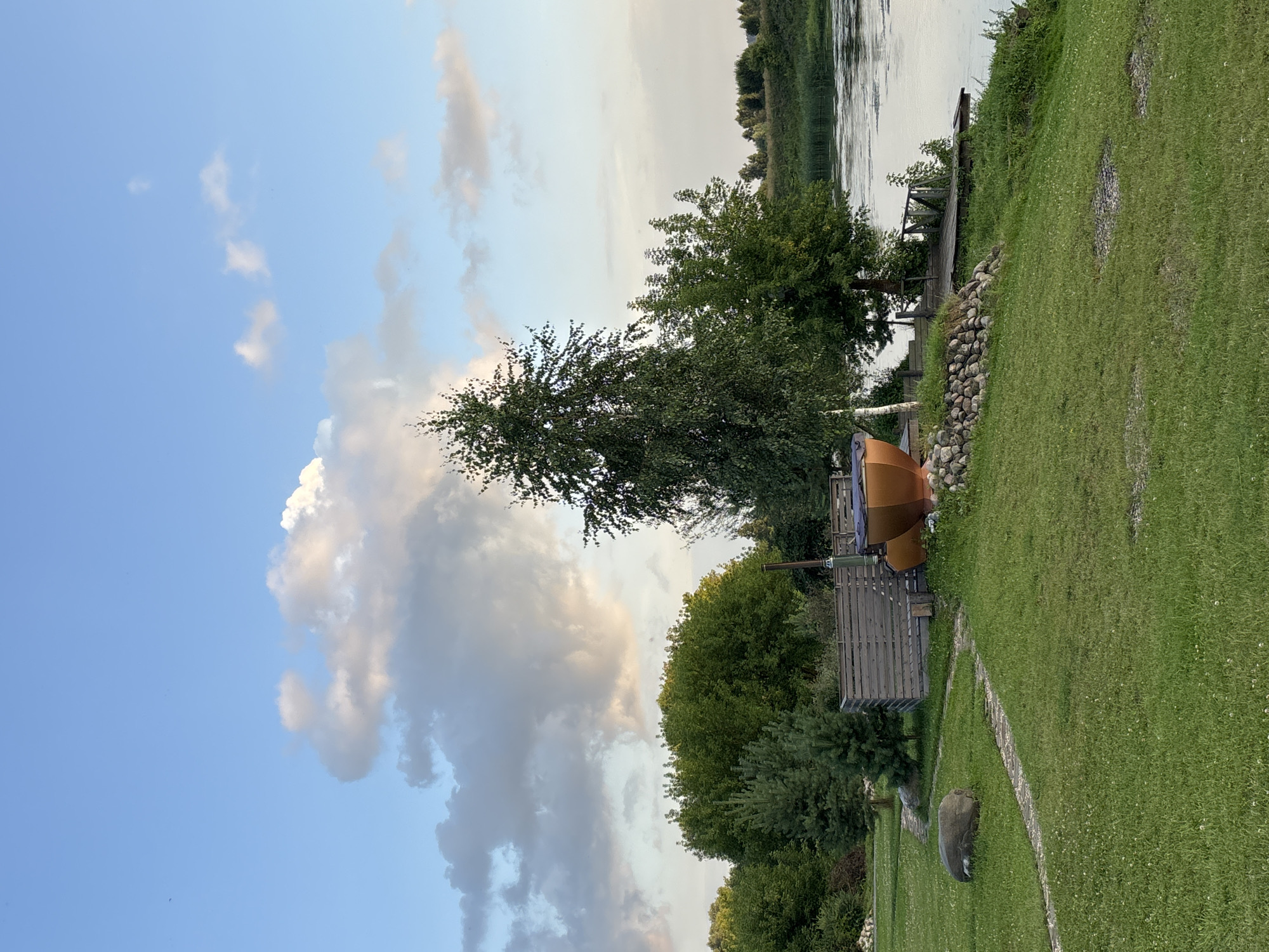 An outdoor scene at dusk features a body of water to the right, reflecting the sky. A wooden dock extends into the water. On the grassy bank, a large, orange, boat-shaped object, possibly a hot tub or sculpture, is visible. A stone wall and a wooden fence are also present in the immediate vicinity. Trees and foliage surround the area, with a tall birch tree dominating the center-left of the frame. The sky is a pale blue with scattered clouds, some of which are illuminated by the setting sun. The ground is covered in lush green grass. The location is a park or recreational area in Zvenigorod, Russia. There are no people or animals visible in the image.