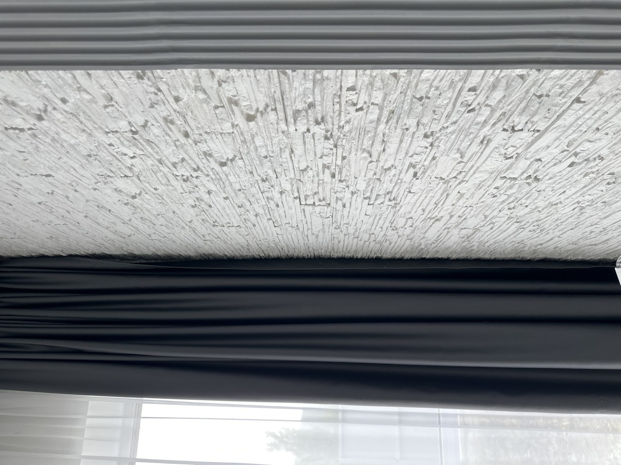 The image captures an upward-angled view of an indoor setting, focusing on a textured ceiling and a window treatment. The top portion of the frame features a white, highly textured surface, resembling a very coarse popcorn ceiling or a decorative plaster finish with numerous small, irregular projections. Directly above this textured area, a gray horizontal element with distinct parallel ridges is visible, possibly a ceiling trim or a valance.
Below the textured white ceiling, a dark, likely black or very dark gray, fabric material is present. This material is arranged in soft, horizontal folds, strongly suggesting a window covering such as a Roman blind or a roller shade that is partially unrolled.
At the bottom of the image, a window is partially visible. It features white horizontal blinds, which appear to be in an open or partially open position, allowing ambient light to pass through. Beyond the blinds, out-of-focus elements of an outdoor scene are discernible, including what appears to be foliage or trees under bright daylight conditions.
The perspective is from below, looking up towards the ceiling and the upper part of the window. The scene depicts a static interior, with no people or discernible activity. There is no visible text in the image. The time of day is clearly daytime, given the light filtering through the window.