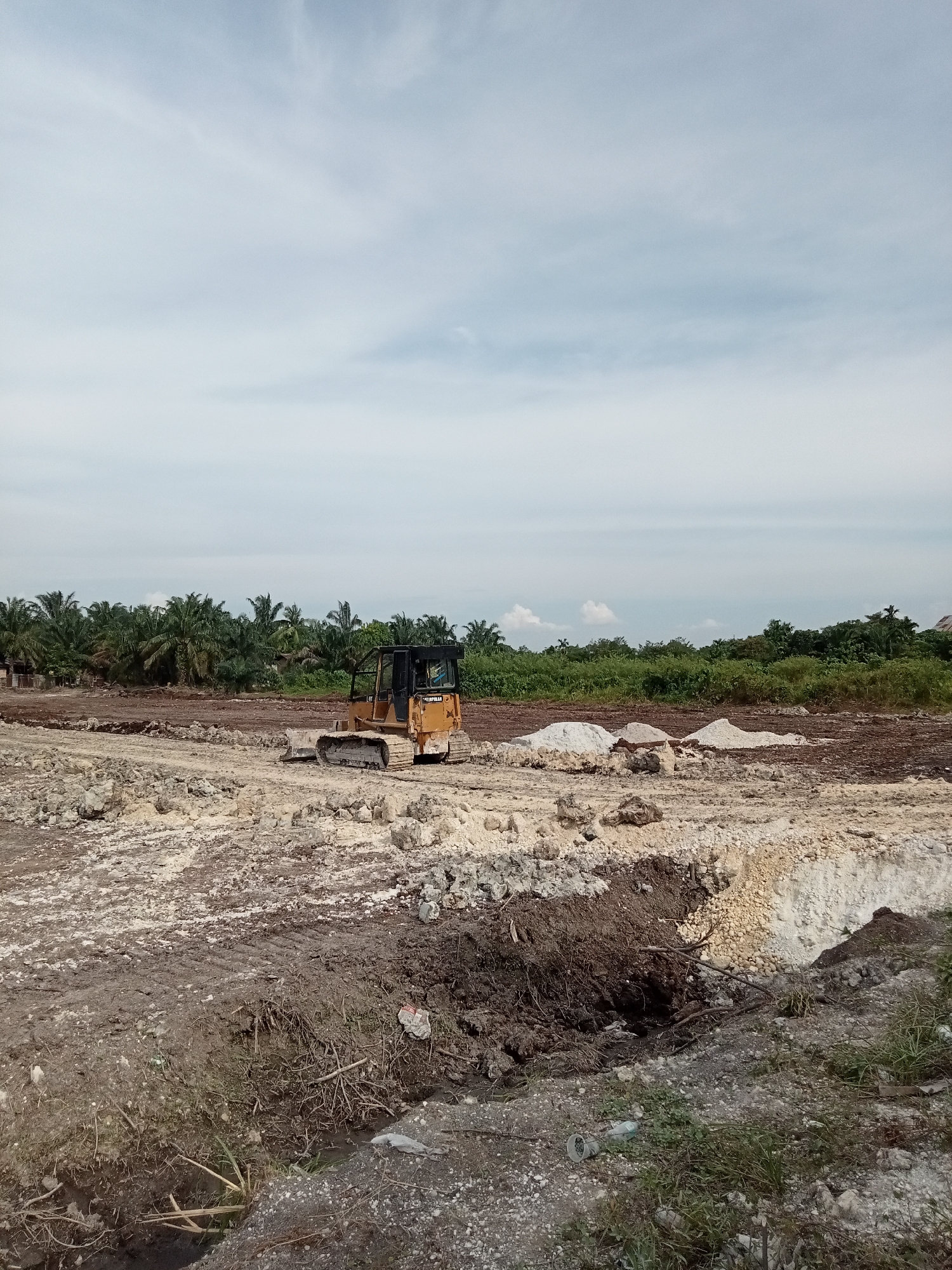 The image depicts a construction site in Teluk Kenidai, Indonesia, under a partly cloudy sky. A yellow bulldozer, identified by the "CAT" logo on its side, is positioned on the uneven, dirt terrain. The ground is a mix of loose soil, gravel, and some debris. In the background, a line of palm trees and dense green vegetation is visible, indicating a tropical environment. The site appears to be in the early stages of development, with cleared land and piles of excavated material. No people are visible in the image.