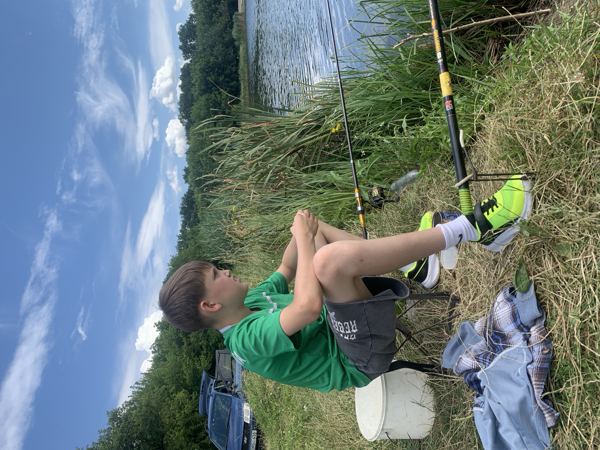 A young boy with short brown hair, wearing a green t-shirt and grey shorts, is sitting on a foldable stool next to a white bucket. He is facing to the right, looking towards a body of water. Two fishing rods are set up in front of him, positioned near the water's edge. In the background, a blue car with its trunk open is parked on a grassy slope, with a line of trees behind it. The sky is blue with scattered white clouds, and tall reeds and grass grow along the bank of the water. The scene appears to be a fishing trip in a natural setting.