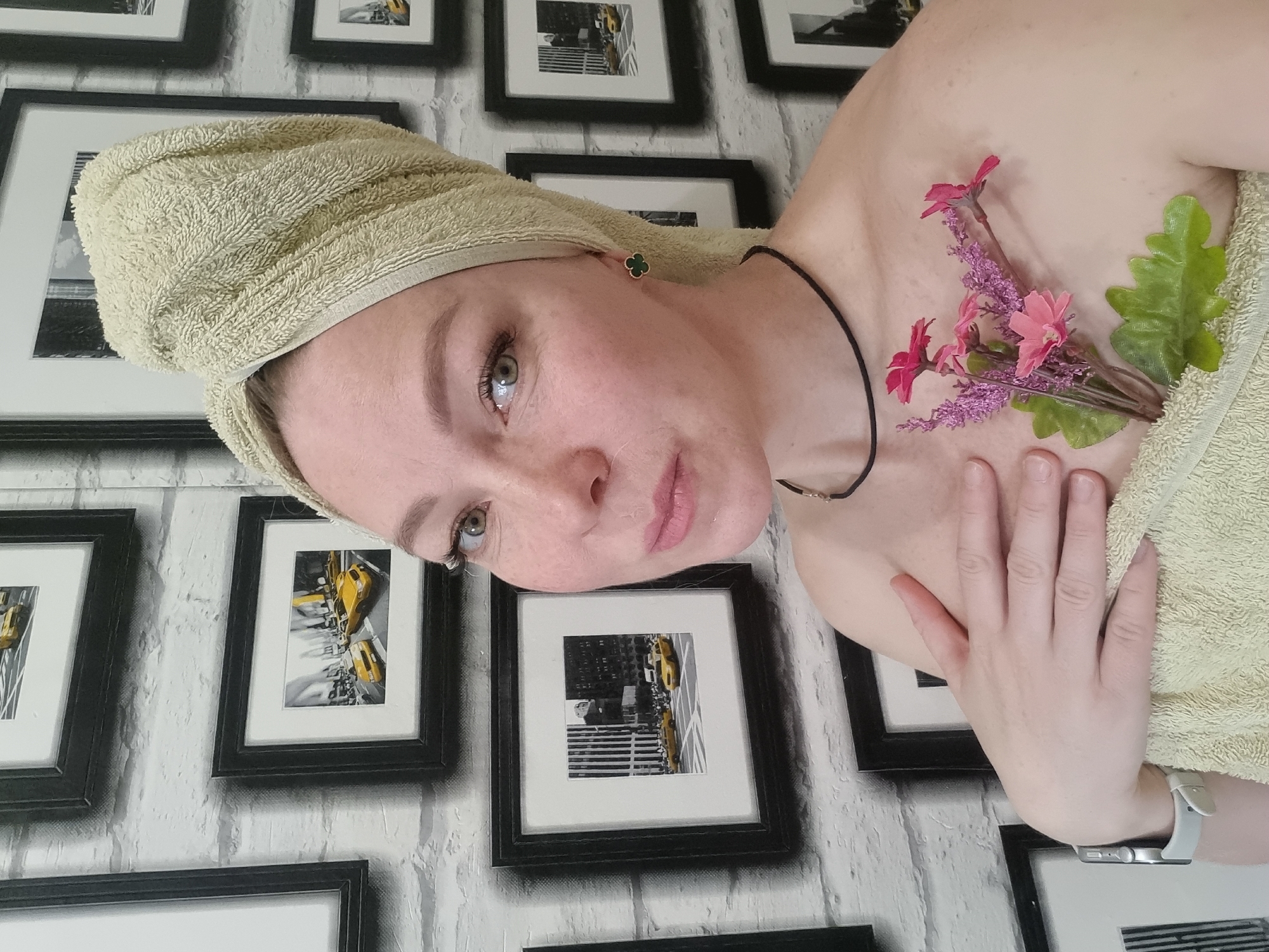 A woman is shown from the chest up, angled slightly to the left. She has a towel wrapped around her head and another draped across her upper body, partially obscuring her shoulder. A small bouquet of pink and purple flowers, along with a green leaf, is positioned near her collarbone. The woman wears a delicate black choker necklace and a small, clover-shaped earring. Her expression is serene, with light pink lipstick and a faint smile. Her arm is across her body, with a watch visible on her wrist. The background features a white brick-patterned wall adorned with framed black and white photographs that seem to depict scenes from New York City, including yellow taxi cabs. There is no indication of Chaikovsky, Russia.