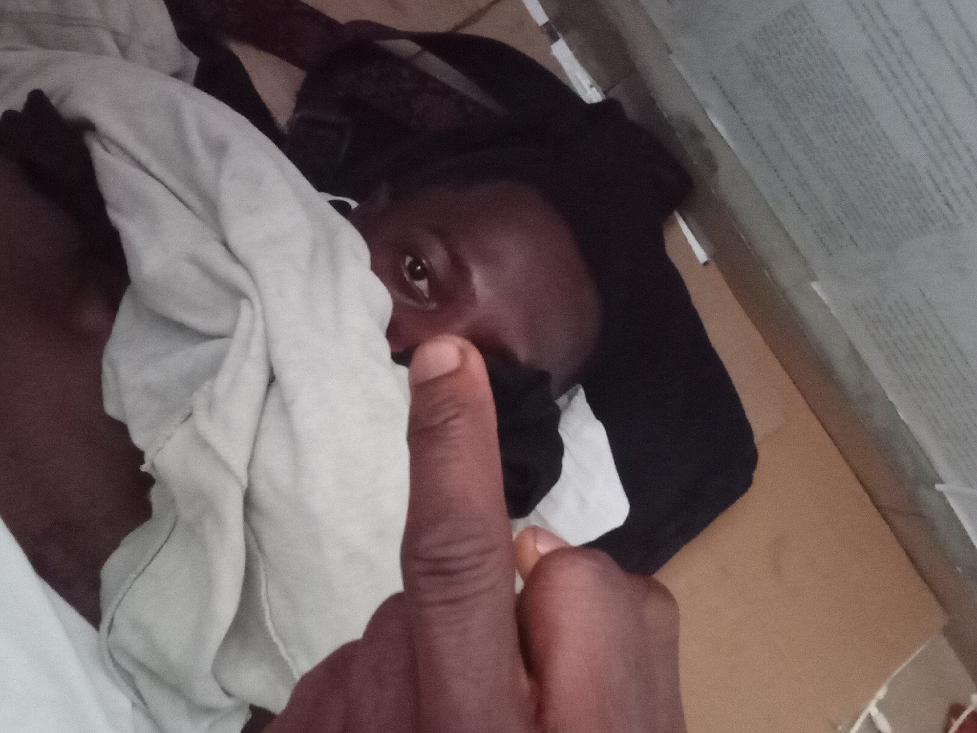 A black man is lying down, partially covered by a grey/white blanket or sheet, on a surface appearing to be cardboard or a similar material. The man is looking at the camera with one eye visible. He is holding up his middle finger. The environment is indoors, with an unidentifiable object or material visible in the upper right corner. The location of Akwete, Nigeria, is not discernible from the image.