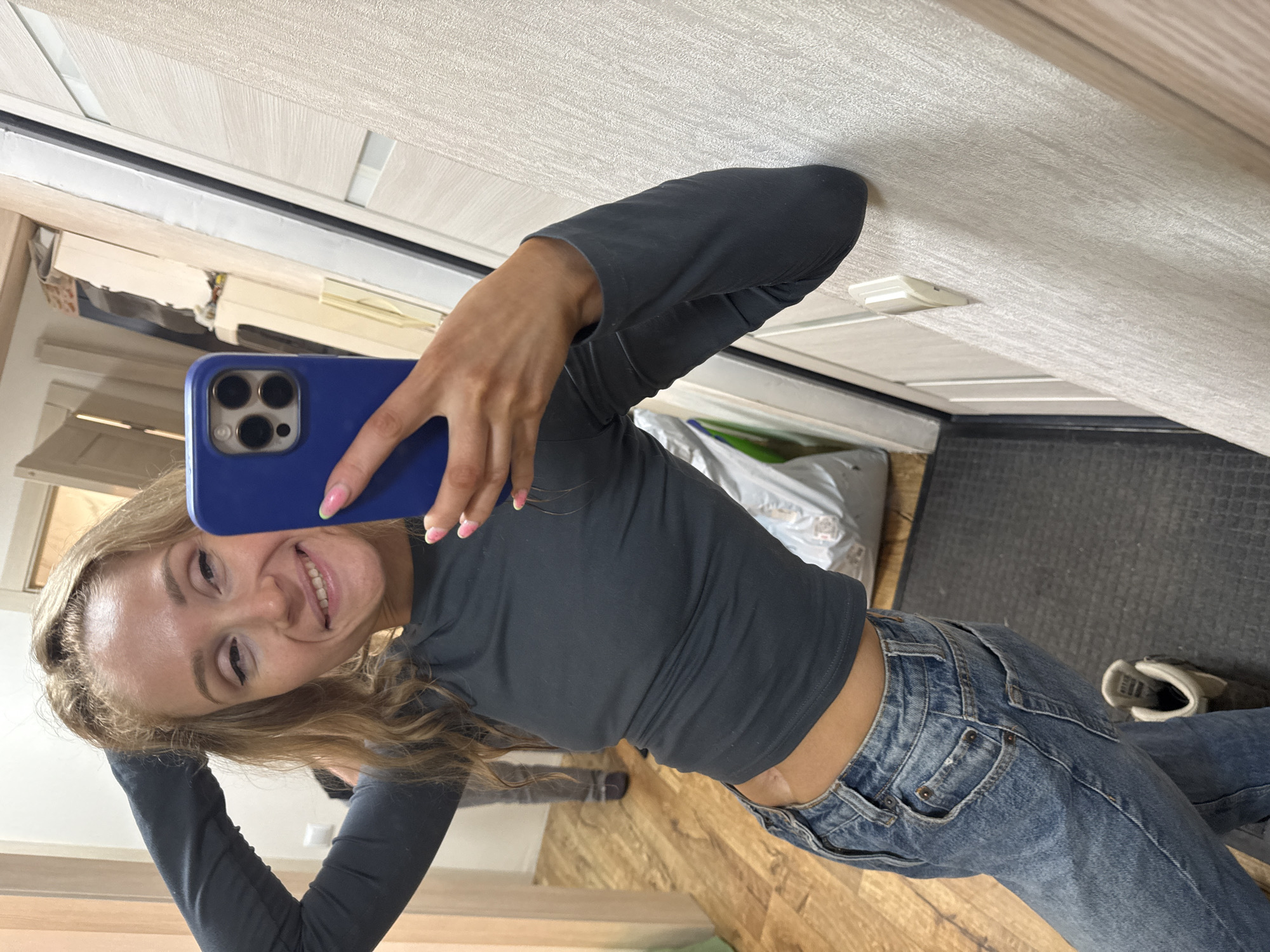 A fair-skinned woman with long blonde hair is taking a selfie in what appears to be an indoor setting. She is wearing a gray long-sleeved shirt and blue jeans. Her phone, encased in a blue cover, is prominently displayed in her hand.
The background shows interior elements like a door, wall, and flooring. Based on the architectural details of the building, flooring material and wall paint, the setting may be a residential space located in Perm, Russia. Other objects in the background include shoes and a plastic bag.