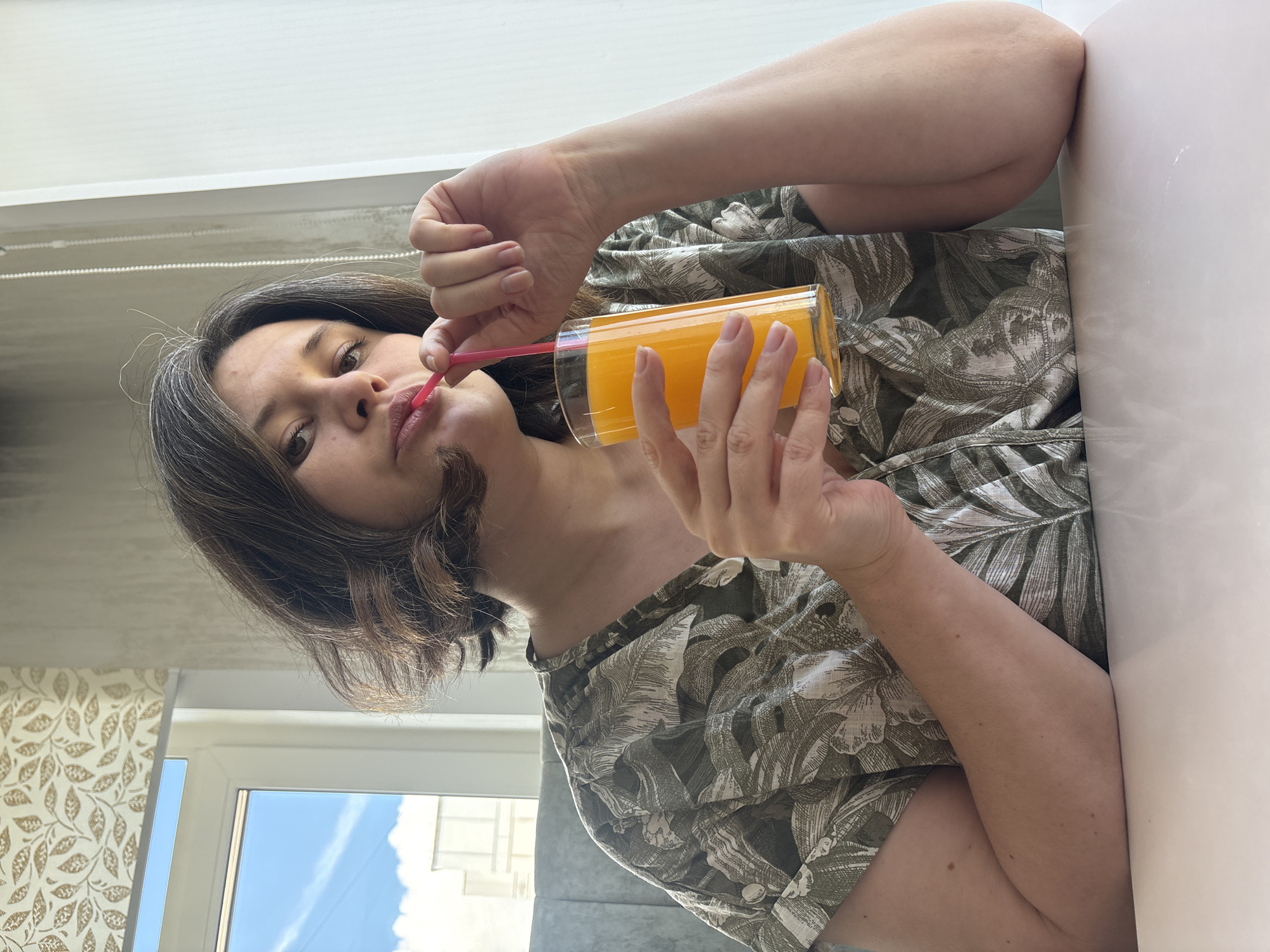 A woman is drinking orange juice through a pink straw. She is holding a glass of orange juice with her right hand. She is wearing a green and white leafy-patterned short-sleeved shirt. Her hair is dark with graying strands. The location appears to be indoors near a window with a view of a blue sky with white clouds. The wallpaper has a leaf pattern. It is possibly Chaikovsky, Russia.