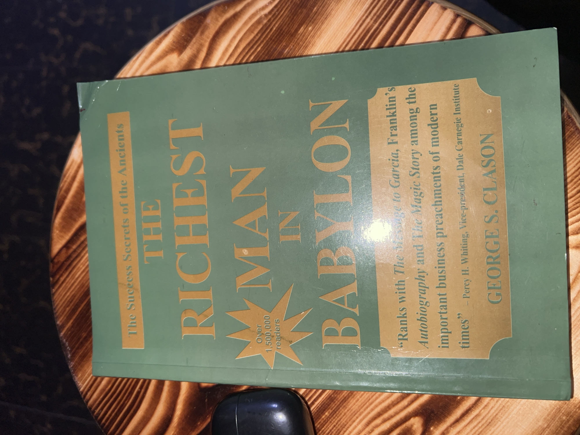The image shows the book "The Richest Man in Babylon" by George S. Clason, lying on a circular wooden surface with a wood-grain pattern. The book's cover is a muted green with gold lettering. At the top of the cover, it reads "The Success Secrets of the Ancients". Below this, the title is prominently displayed: "THE RICHEST MAN IN BABYLON". To the left of "MAN", there is a gold starburst shape with the text "Over 1,500,000 readers". At the bottom of the cover, within a gold-bordered rectangle, is a quote that reads: "“Ranks with The Message to Garcia, Franklin's Autobiography and The Magic Story among the important business preac ments of modern times” - Percy H. Whiting, Vice-President, Dale Carnegie Institute". Below this quote, the author's name, "GEORGE S. CLASON", is printed in gold letters. A black earbud case is partially visible on the left side of the image, resting on the wooden surface next to the book. The lighting in the image is dim, creating shadows and highlighting the textures of the wood and the book cover. The background is dark and out of focus. There are no people visible in the image, and no specific details suggest a location within the city of Lagos, Nigeria.