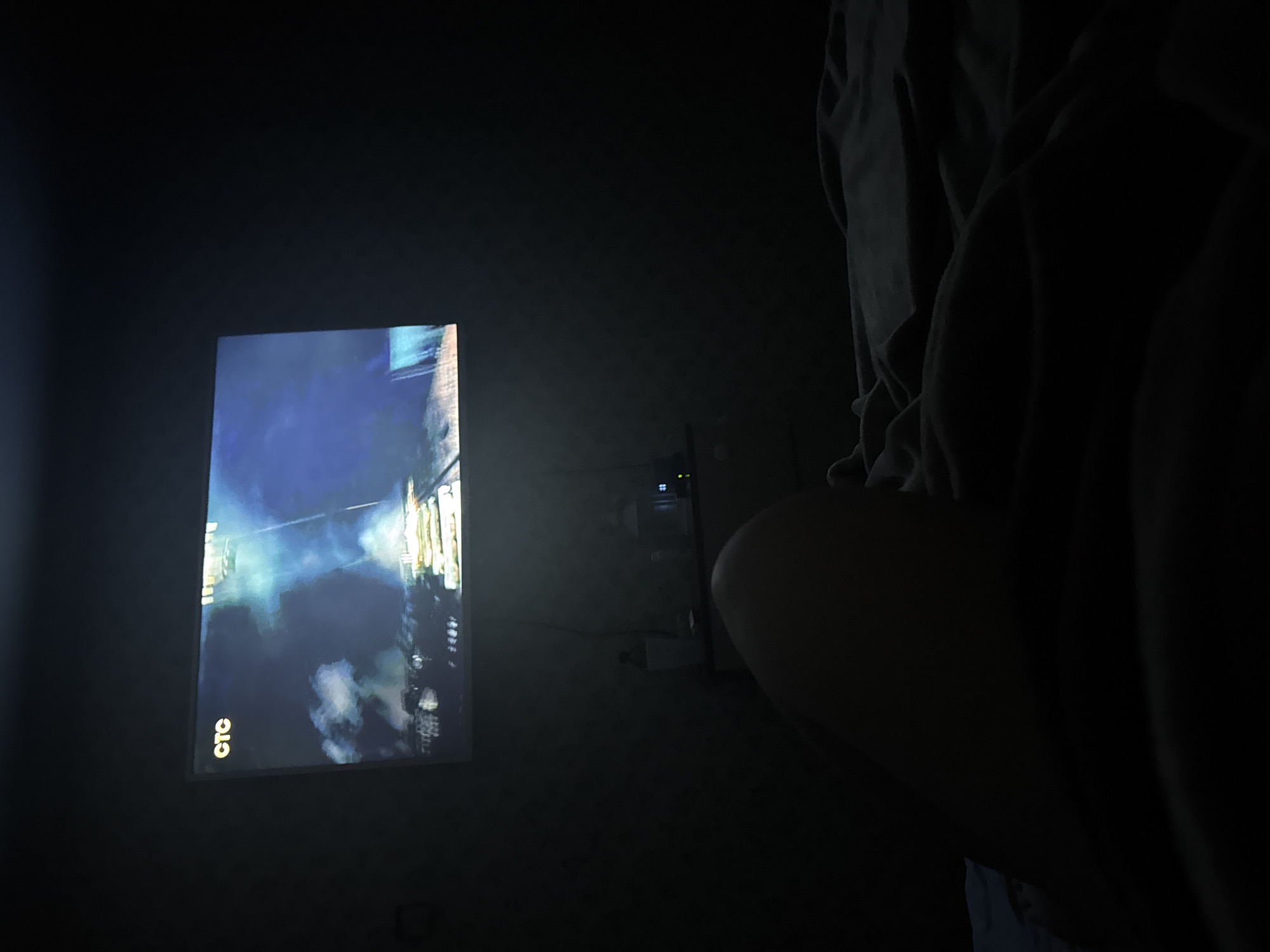 A dimly lit interior features a television screen projecting an image of a cityscape at night. The screen's bottom left corner displays the word "CTC" in Cyrillic script. To the right of the screen, a blurred figure in a hooded sweatshirt is partially visible. Power outlets and cords are faintly discernible on the wall between the screen and the figure. The location cannot be determined from the image.