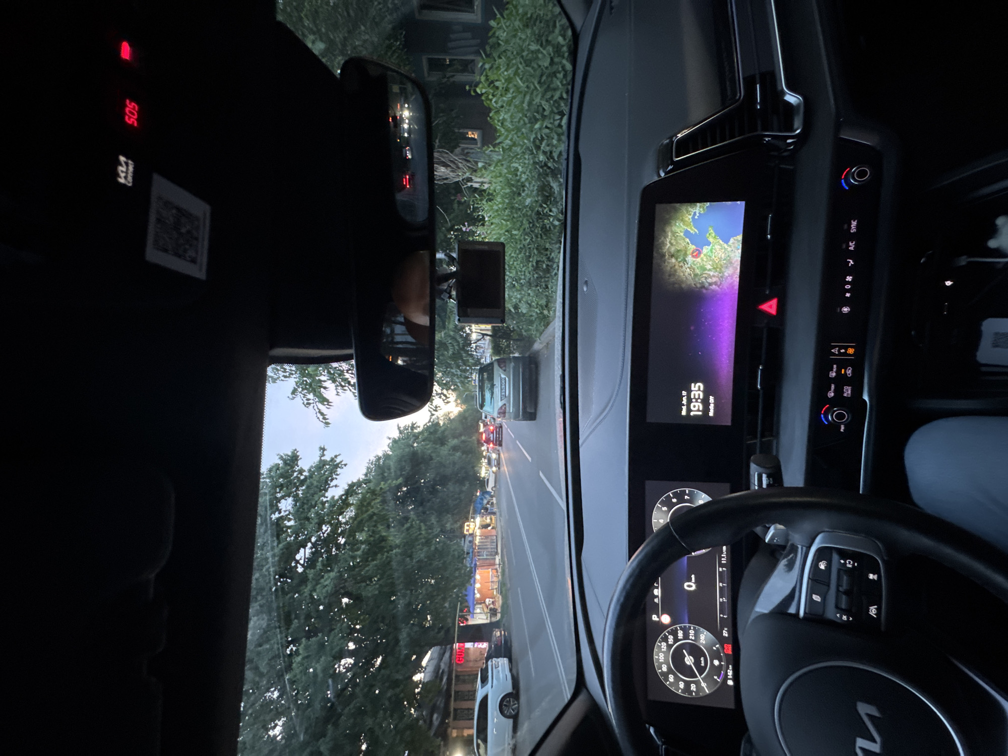 The image shows the interior of a car, likely a Kia, as indicated by the "Kia Connect" logo and the "Ki" logo on the steering wheel. The driver's perspective is evident, looking out through the windshield. The scene is set in Almaty, Kazakhstan, on a street lined with trees and buildings. The time is 19:35 on Wednesday, June 17th, suggesting it's early evening. The weather appears to be clear as there is visible sky.
The car's interior features a modern dashboard with a large digital display showing navigation information with a map and possibly media details. A red emergency light labeled "SOS" is visible above the rearview mirror. The steering wheel is equipped with various controls. Outside, the street is moderately busy with other vehicles, and there are some market stalls or shops visible on the right side of the road, illuminated by lights. The overall impression is of a typical urban scene in Almaty during the early evening commute.