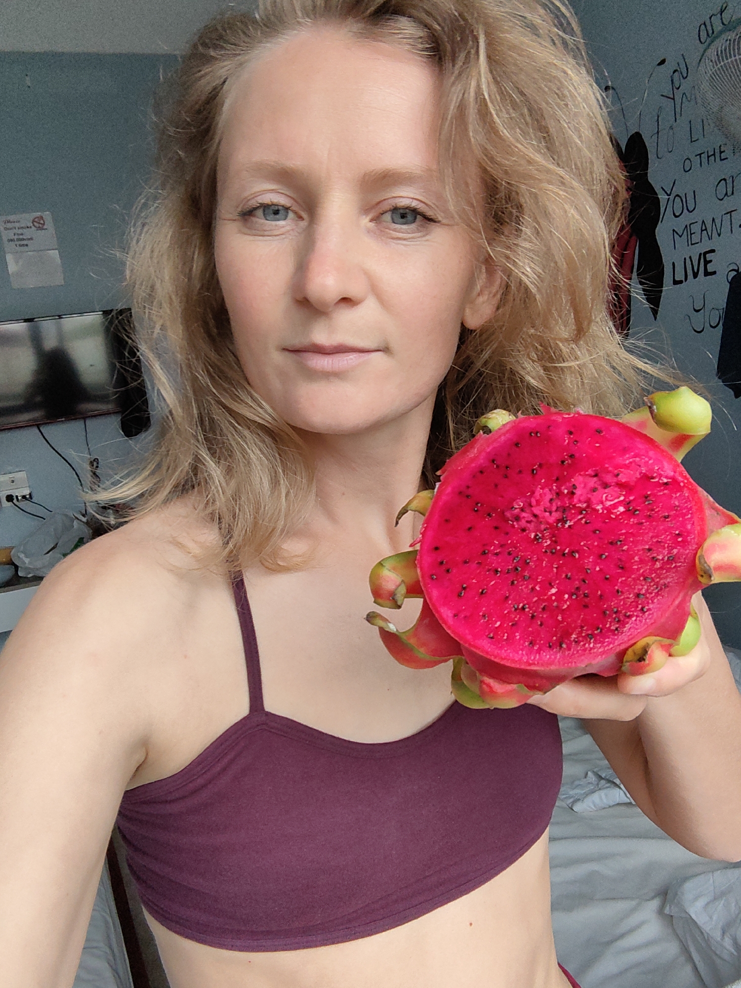 A woman in a maroon sports bra holds a halved, vibrant magenta dragon fruit in her right hand. She has fair skin, blue eyes, and blonde, wavy hair. The background includes a partially visible television, a wall with a "no smoking" sign, a fan, and a wall with motivational text. A bed with white linens is also visible. It's likely an interior shot taken in a residential setting. The location is within Bai Chay Ward, Vietnam.