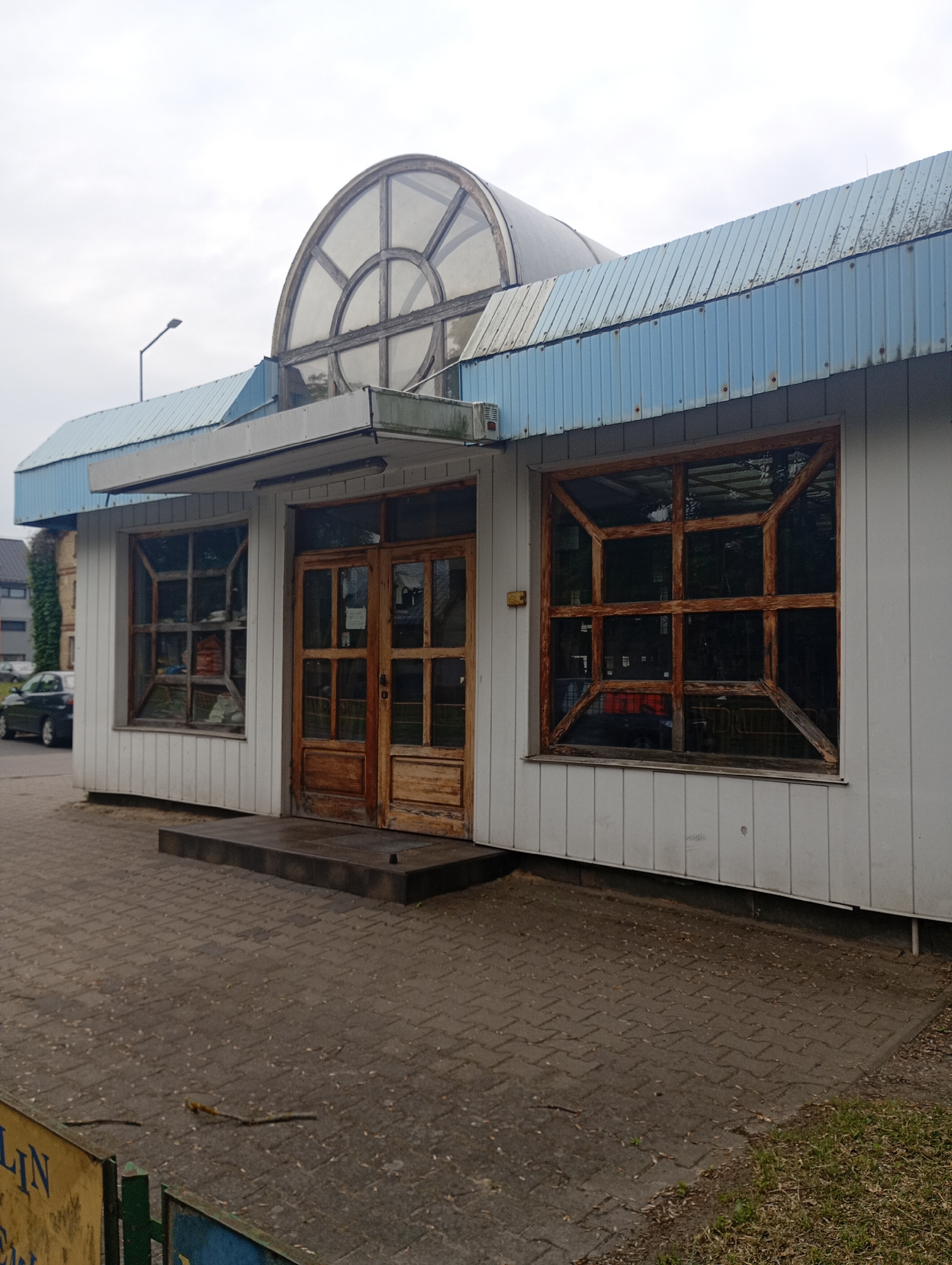 This is a medium-shot, eye-level, outdoor photograph taken in Międzyrzecz, Poland, of the exterior of a single-story building. The building has a white, wood-paneled facade and a light blue, corrugated metal roof. A prominent semicircular, glass-paneled skylight with a wooden frame is situated on the upper portion of the structure. There are two wooden doors with glass panes, centrally located on the front of the building, leading to the interior. To the left of the doors, there is a large, square window with a wooden grid pattern and tinted glass. To the right of the doors, another similar window is visible. The foreground consists of a paved walkway made of interlocking bricks, with some debris and sparse grass on the right side. A dark-colored car is parked on the street to the left of the building. The sky is overcast.