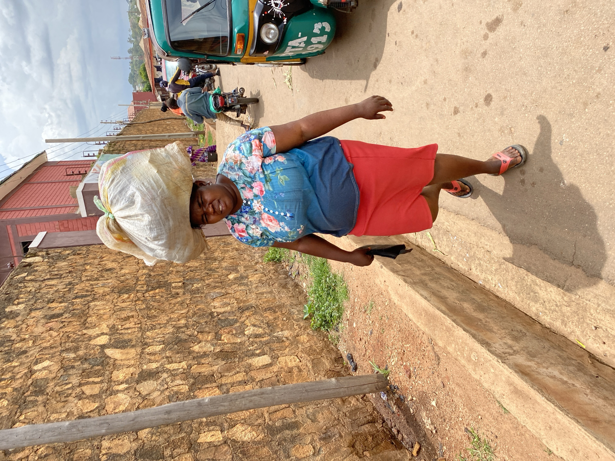 In Bukuru, Nigeria, a woman with a large, lumpy white sack balanced on her head walks down a paved road. She is wearing a blue and floral print short-sleeved shirt and a bright red skirt. Her right hand clutches a black item, possibly a cloth. She is wearing orange flip-flops. To her left is a textured, tan stone wall. A gray wooden pole stands near the wall. A concrete curb runs along the edge of the road, with some sparse green vegetation growing beside it. To her right, a green and yellow motorized tricycle is parked. Several other people are visible in the background, with two on a motorcycle and one walking further down the road. The sky is overcast with scattered clouds.