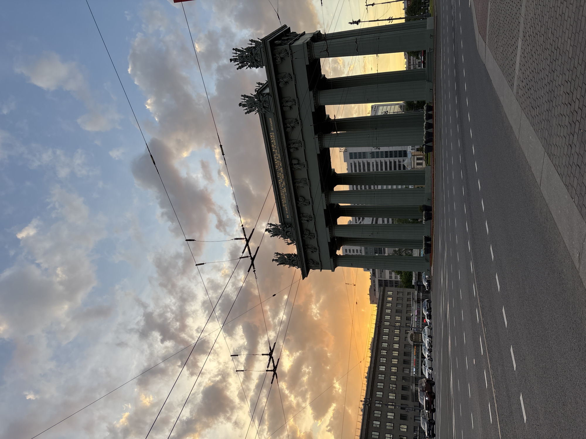 The image displays the monumental Narva Triumphal Arch in Saint Petersburg, Russia, under a dramatic sky, likely at sunrise or sunset. The sky transitions from a soft blue with scattered clouds on the left to a vibrant golden-orange hue near the horizon, casting a warm glow on the scene.
The central subject is the grand classical arch, featuring multiple tall, fluted columns supporting a detailed entablature. The arch is adorned with intricate sculptural reliefs and figures atop its structure. A prominent inscription in Cyrillic is visible on the entablature, reading: "РОССІЙСКИМЪ ВОЙСКАМЪ ПОБѣДИТЕЛЯМЪ ПОСЛѢДНѢЙ ОТЕЧЕСТВЕННОЙ ВОЙНЫ 1812 ГОДА И ВЪ ПАМЯТЬ ПОХОДА ВОЗВРАЩЕНИЯ ИХЪ ИЗЪ ПАРИЖА ВЪ 1814 ГОДУ" (To the victorious Russian troops of the last Patriotic War of 1812, and in memory of their campaign and return from Paris in 1814).
In the foreground, a wide, empty asphalt road with clear white dashed lane markings stretches towards the viewer and into the distance. To the far right, a section of patterned pavement or cobblestones is visible. Numerous black utility lines, likely for trolleybuses or trams, crisscross the sky, adding a contemporary urban layer to the historic architecture.
Beyond the arch, modern multi-story buildings can be seen, some reflecting the golden light of the sky. Further down the road, additional buildings line the street, with several cars parked alongside the curb. The street appears largely deserted, contributing to a serene atmosphere. The overall scene combines historical grandeur with urban elements under the striking light of dawn or dusk.