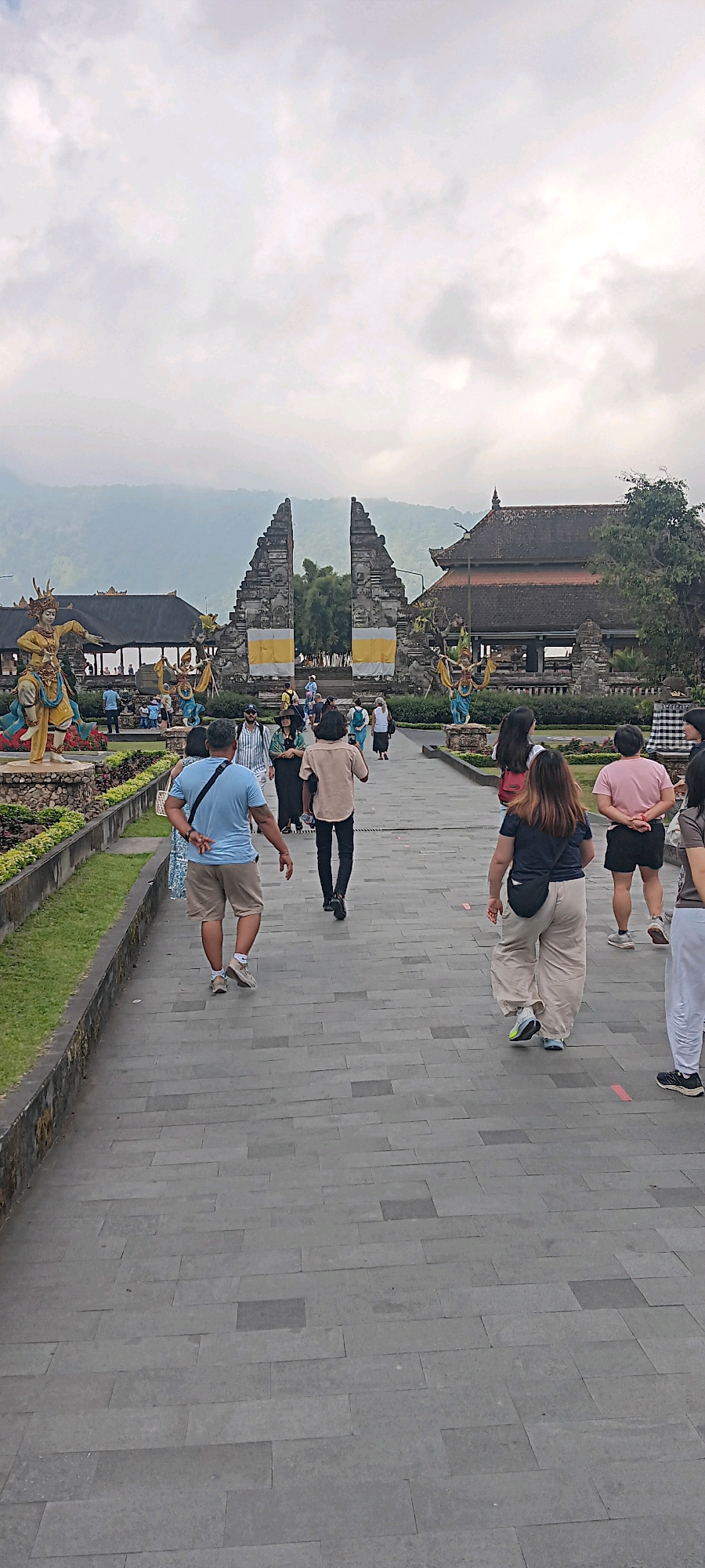 In the city of Baturiti, Indonesia, a group of tourists walk along a paved pathway towards a large stone gate. The gate is flanked by traditional Balinese statues dressed in ornate costumes. Beyond the gate, a temple building with a dark, pitched roof is visible. The sky is overcast with clouds. The pathway is lined with greenery and decorative stone borders. Several individuals are captured mid-stride, some with their backs to the camera. One person in the foreground wears a light blue t-shirt and khaki shorts. Another person further ahead is dressed in a light brown collared shirt and dark pants. On the right side of the pathway, a person in a pink t-shirt and black shorts is visible, and next to them, someone in white pants. The overall scene depicts a cultural landmark and a tourist attraction.