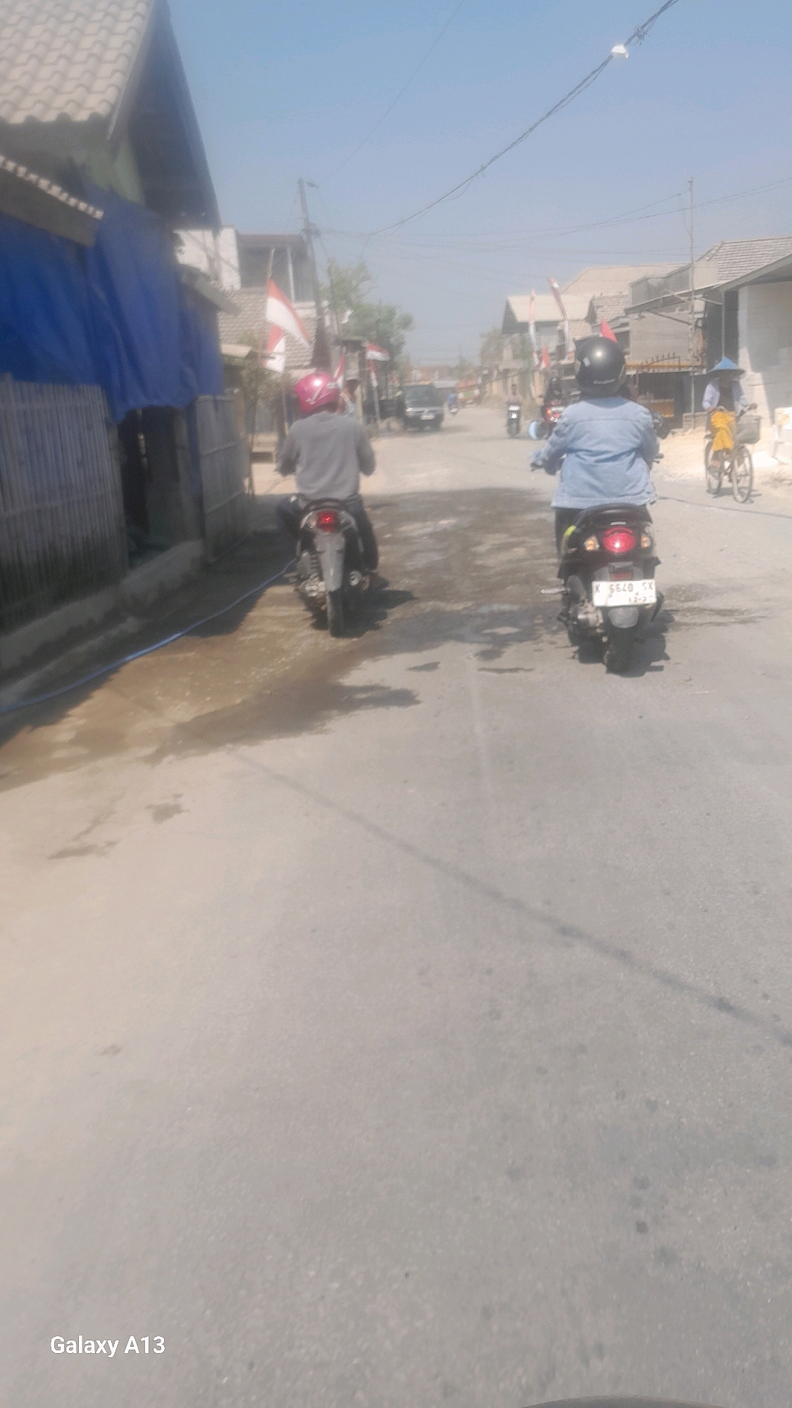 Two people are riding motorcycles on a street in Ngeseng, Indonesia, under a clear sky. The motorcycle in the foreground has a license plate reading "K 5540 SK". In the background, a person is riding a bicycle. The street appears to be paved with asphalt and has some wet patches. Buildings line the street, some with corrugated metal roofs. Flags are visible, suggesting a local celebration or event. The overall scene depicts a typical day in a residential area.