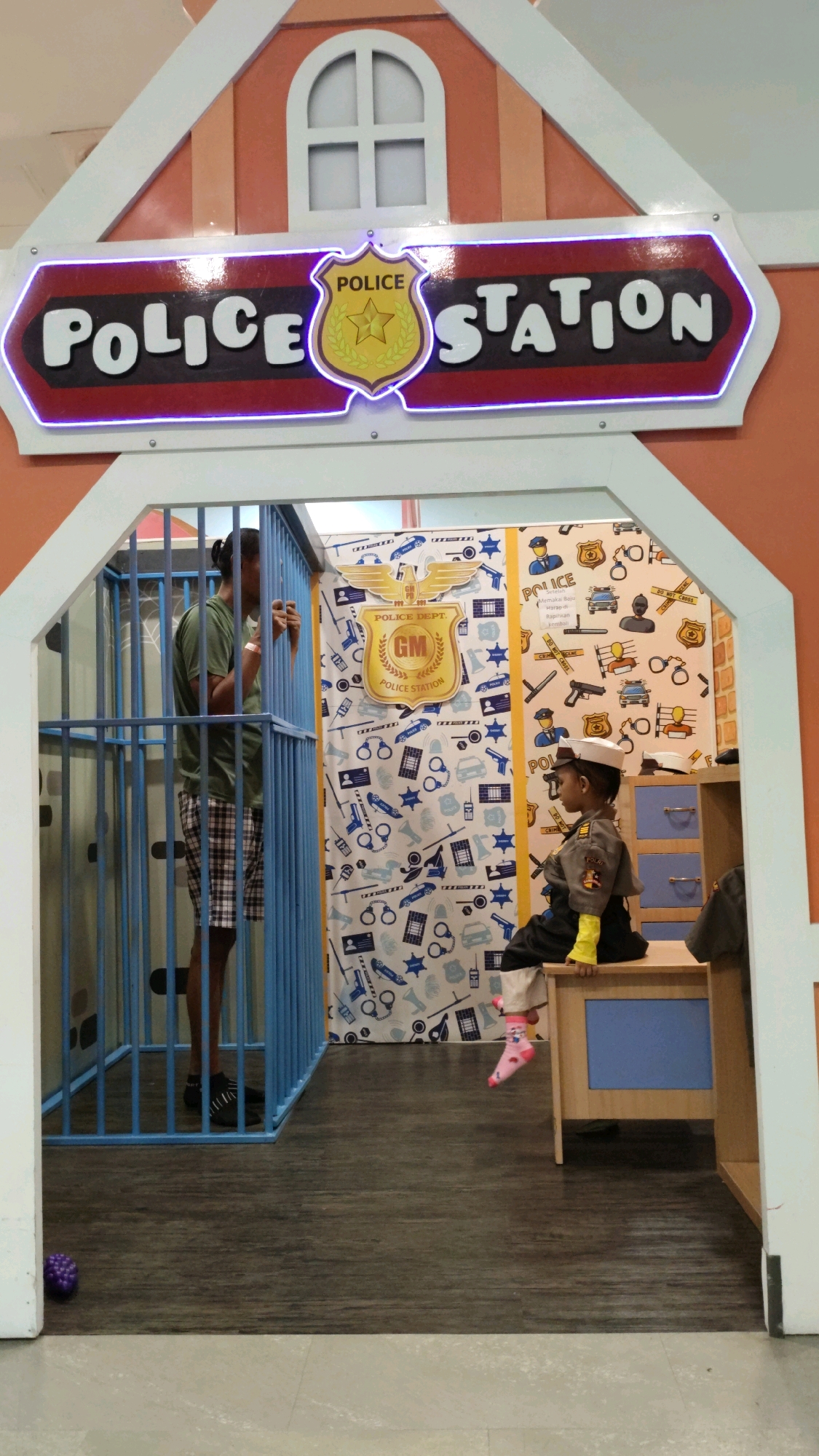A man stands inside a blue-barred jail cell, leaning forward with his hands on the bars. He is wearing a dark green t-shirt and plaid shorts. In front of the jail cell, a young child, dressed as a police officer with a hat, jacket, and yellow sleeves, sits on a wooden desk with blue drawers. The child is wearing pink socks with cartoon characters. Behind the child is a wall decorated with police-themed patterns. Above the jail cell is a sign that reads "POLICE STATION" in white lettering with a neon purple outline. The scene appears to be a play area, possibly in a museum or children's entertainment center. The flooring is a dark wood grain laminate. A purple ball is visible on the floor to the left of the jail cell. The overall environment is brightly lit.