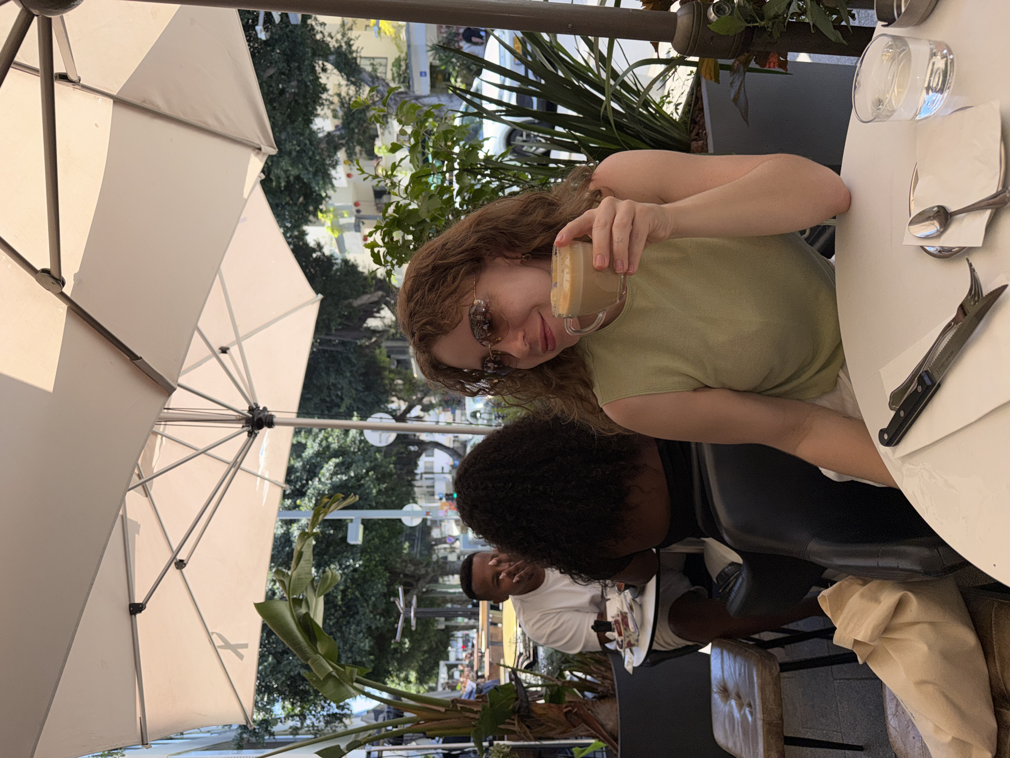The image depicts an outdoor café setting, likely in Tel Aviv, Israel, given the context. A fair-skinned woman with red hair is the focal point, holding up a small glass of coffee or a similar beverage. She is wearing sunglasses and a light green sleeveless top. Behind her is a black woman and an African American man in a white t-shirt sits at a table. There are tables with utensils and glassware on a white tablecloth. Above, a large beige umbrella provides shade, and trees and greenery are visible in the background, suggesting an urban environment. The overall atmosphere is that of a relaxed, outdoor dining experience.
