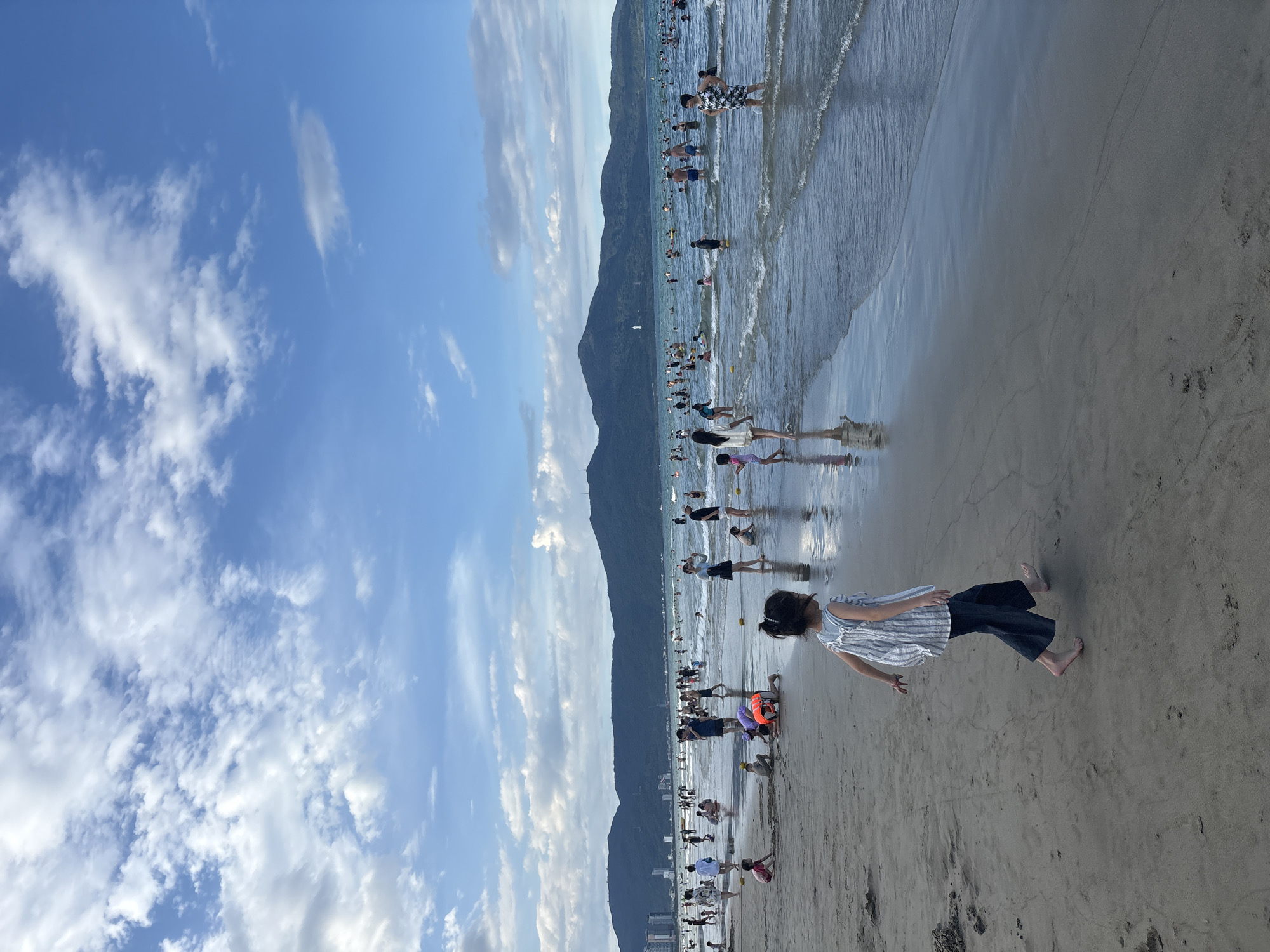 This is a vertical, low-angle, full-shot photograph of a crowded beach in Đà Nẵng, Vietnam. The scene depicts a lively beach with numerous people enjoying the water and sand under a bright, cloudy sky.
The primary subjects are the many people spread across the beach and wading in the shallow water. There are adults and children visible, some swimming, others walking along the shoreline, and some sunbathing or relaxing on the sand. The setting is a sandy beach with gentle waves lapping at the shore. In the background, a lush, green mountain range dominates the horizon, providing a scenic backdrop. The sky is a vibrant blue, dotted with fluffy white clouds that suggest a pleasant, possibly late afternoon, time of day. The reflections of the people and the sky are visible on the wet sand near the water's edge, adding depth to the image.
The activity taking place is recreational beach-going, with people engaged in swimming, wading, and enjoying the outdoors. The atmosphere appears to be cheerful and relaxed, with a general sense of summer enjoyment.
Notable details include the clear blue sky with scattered clouds, the imposing mountain range, and the large number of people enjoying the beach. The wet sand creates a reflective surface. The image is shot vertically, giving prominence to the sky and the expanse of the beach. There is no visible text in the image.
