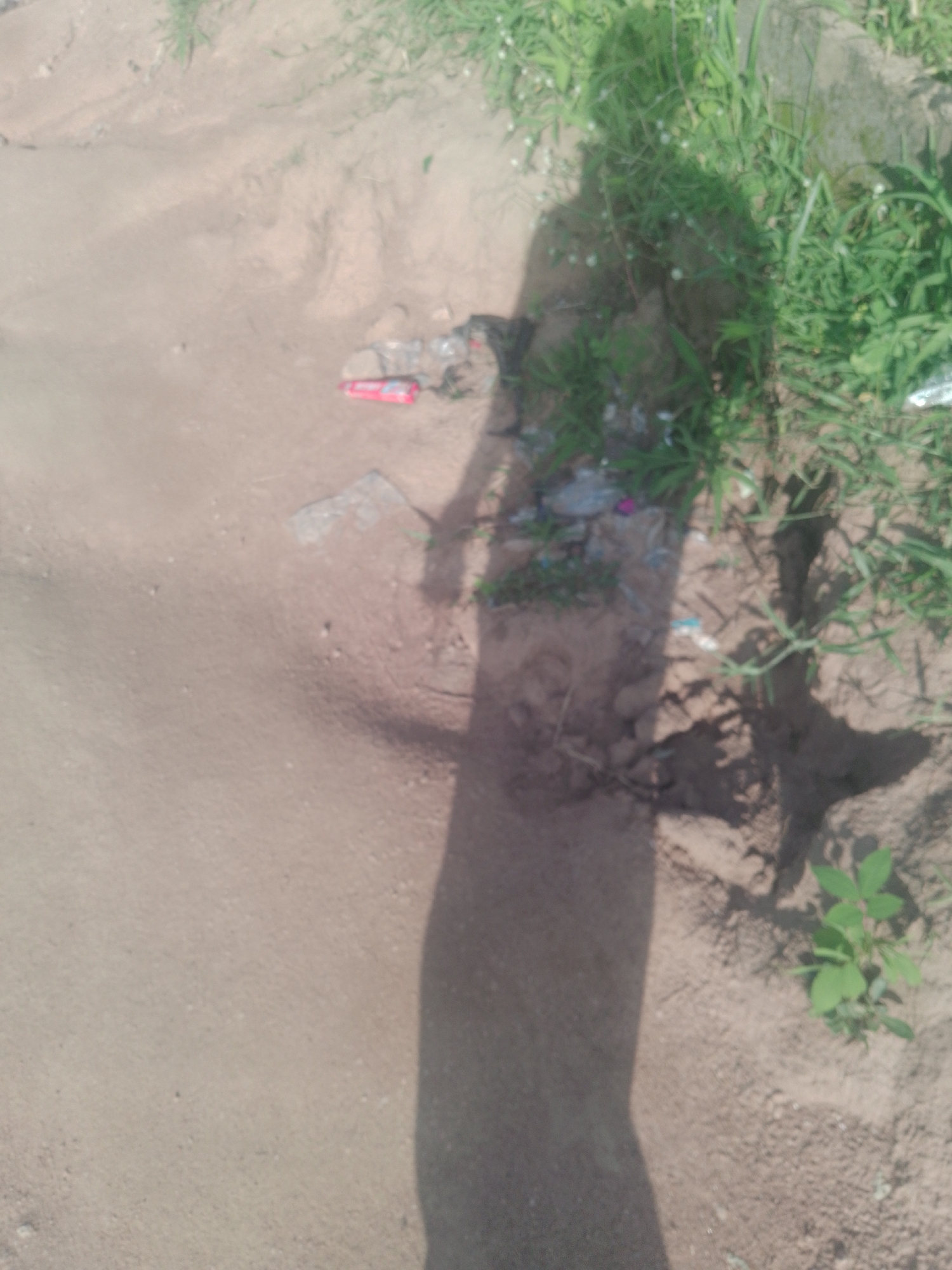 The image shows a shadow of a person standing in an outdoor environment with sparse vegetation and sandy soil. There is some litter visible on the ground, including a red object that appears to be a cigarette lighter and some plastic fragments. The scene is likely located in Jalingo, Nigeria, given the context of the request, but there are no specific landmarks or identifying features to confirm this location within the image itself. The lighting suggests it is daytime.