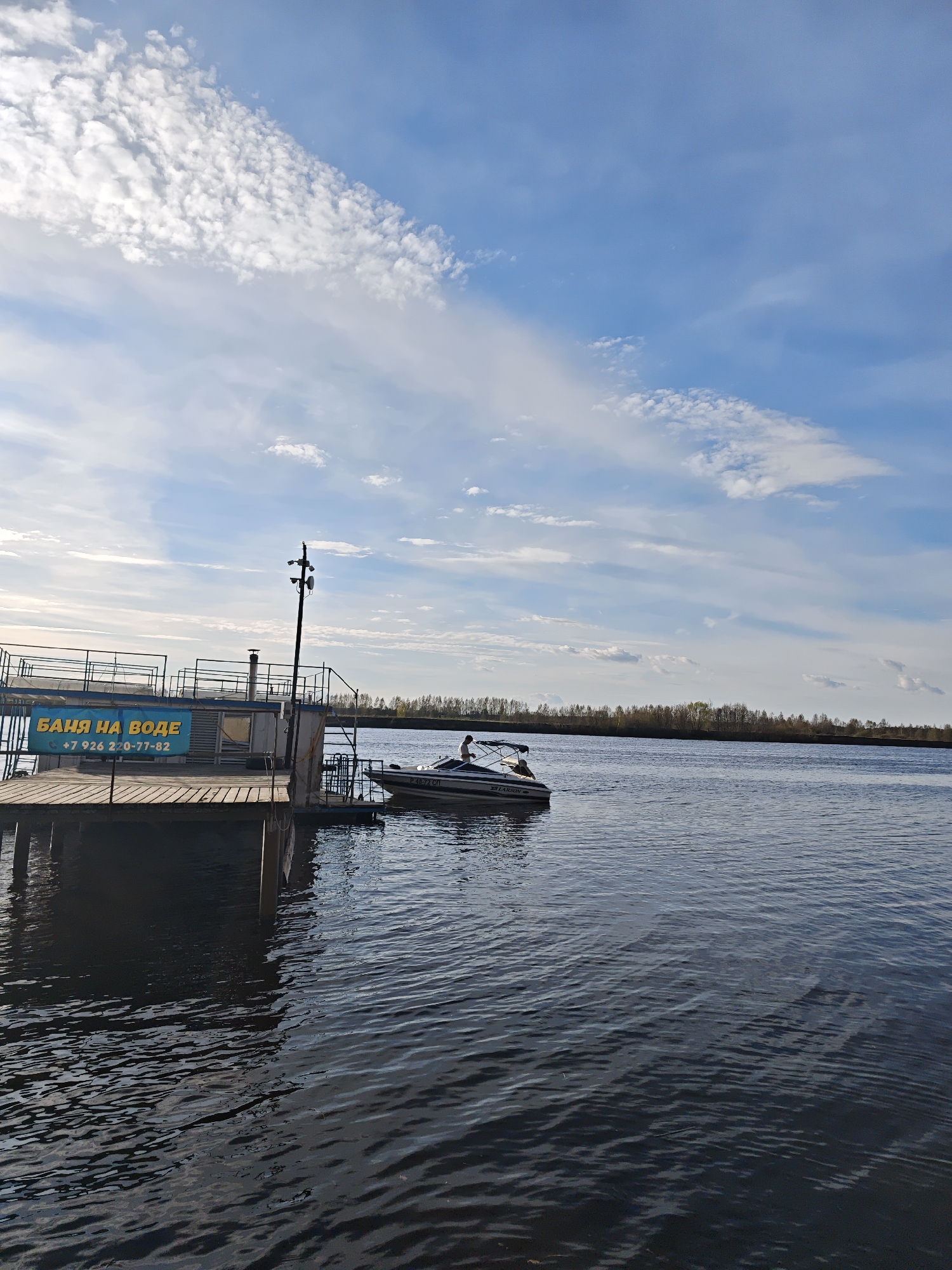 The image shows a waterfront scene with a wooden pier extending into a body of water. Attached to the pier is a structure with the Russian text "БАНЯ НА ВОДЕ" and a phone number "+7 926 220-77-82" displayed on it. A boat, identified as a LARSON, is docked next to the pier, with a person visible inside. A row of trees lines the horizon on the opposite side of the water. The sky is blue with scattered clouds.