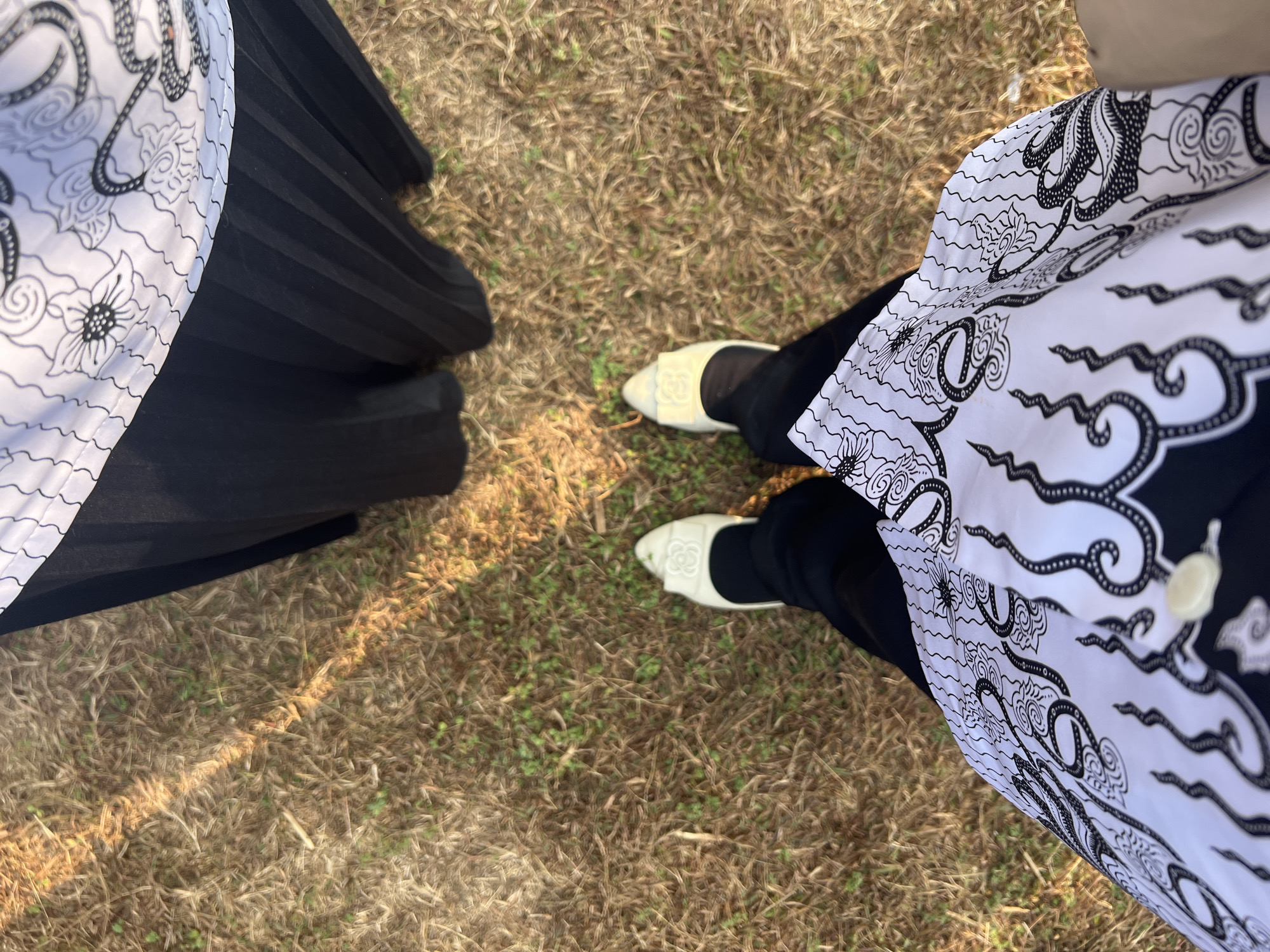 A first-person perspective shot captures two individuals standing on dry grass with scattered green patches. Sunlight casts diagonal shadows across the ground. One person wears white slip-on shoes with a decorative rose motif and black pants, paired with a white and black batik patterned top. Their feet are positioned centrally in the frame. Behind them, another person wears a black pleated skirt and a white and black batik top, with their feet not visible. The scene suggests an outdoor setting, possibly in a park or open field within the city of Sukamantri, Indonesia.