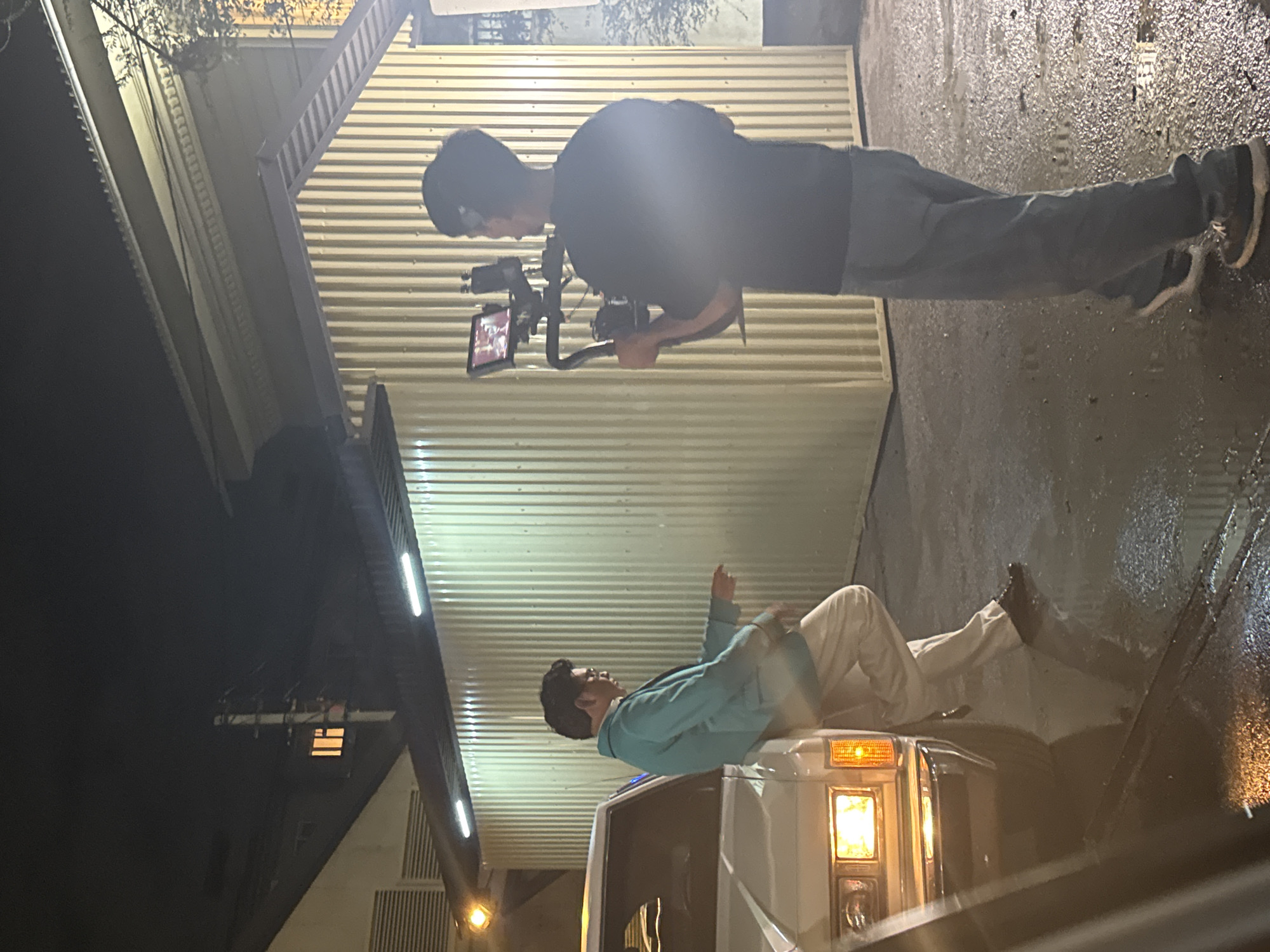 This media file shows two individuals in an outdoor urban setting at night. One person, wearing dark clothing, is operating a video camera on a gimbal. This individual is standing on a wet, paved surface, possibly a street or parking lot, which reflects the ambient light. The background features a building with ribbed siding and some overhead structures, illuminated by artificial lights.
The second person is lying on top of a white vehicle, which appears to be a van or truck. This individual is wearing a light blue jacket and light-colored pants. Their head is propped up, and one hand is raised, as if in conversation or gesturing. The vehicle has its headlights on, casting a warm glow. The ground is wet, indicating recent rain or damp conditions.
The overall mood is somewhat casual and potentially creative, given the presence of video equipment and the posed nature of one of the subjects. The time of day is clearly night, and the weather appears to be wet, adding a reflective quality to the scene. There is no visible text in the media file.
