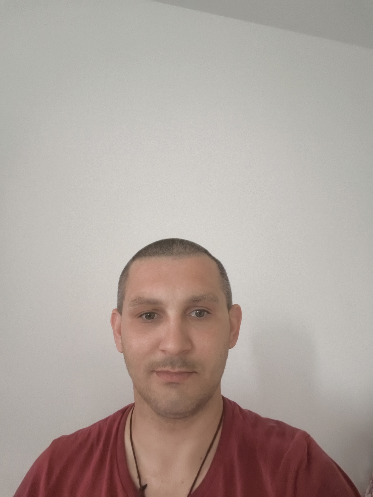 A close-up, waist-up portrait of a light-skinned man with a shaved head and brown eyes is captured against a plain, light-colored background. He is wearing a dark red t-shirt. The man's gaze is directed forward, and he has a neutral expression. A dark cord necklace is visible around his neck. The background suggests an indoor setting, possibly a room with a simple wall. No specific location in Wolgast, Germany is identifiable from the visual information provided in the image.