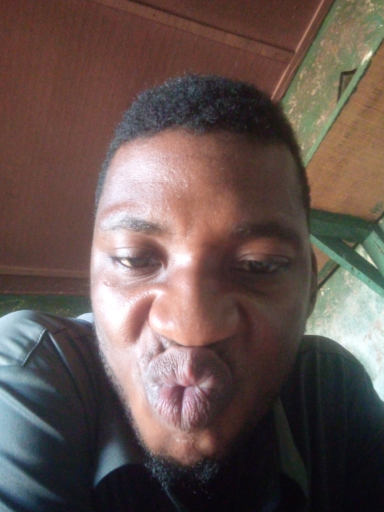 A close-up, slightly low-angle selfie features a person with dark skin, short black curly hair, and a short beard. The person is making a playful "kissing" or "fish lips" expression, with their mouth puckered forward. Their eyes are partially closed and looking downwards, adding to the humorous or introspective mood. Strong lighting illuminates the forehead, nose, and lips, creating highlights. The person is wearing a dark, possibly black or dark grey, collared shirt, visible on the left side of the frame.
The background is somewhat out of focus but provides clues about the setting. On the left and top, there is a reddish-brown or dark wood surface, possibly a wall or ceiling. On the right side, a green-painted wooden structure is visible, which appears worn or distressed, with patches of lighter color showing through. This structure suggests an indoor or sheltered outdoor setting, possibly a shed, balcony, or a room with rustic elements. No discernible text is visible in the image. The overall impression is a casual, personal, and expressive photograph.