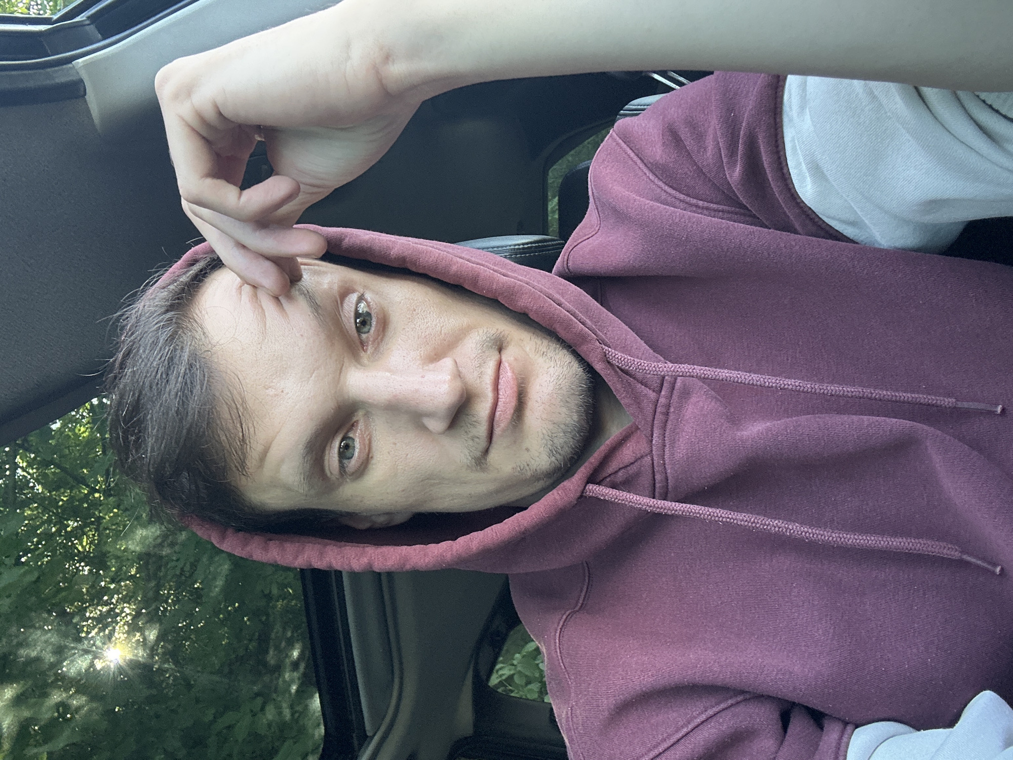 A man wearing a maroon hoodie and a light gray long-sleeved shirt is inside a vehicle, the top of which appears to be a sunroof open to a canopy of green leaves and sunlight. The man is looking directly at the camera, with his right hand resting on his forehead. The interior of the vehicle, including the seats and dashboard, is visible. The background outside the vehicle shows a bright sun shining through dense foliage. The image appears to have been taken outdoors on a sunny day.