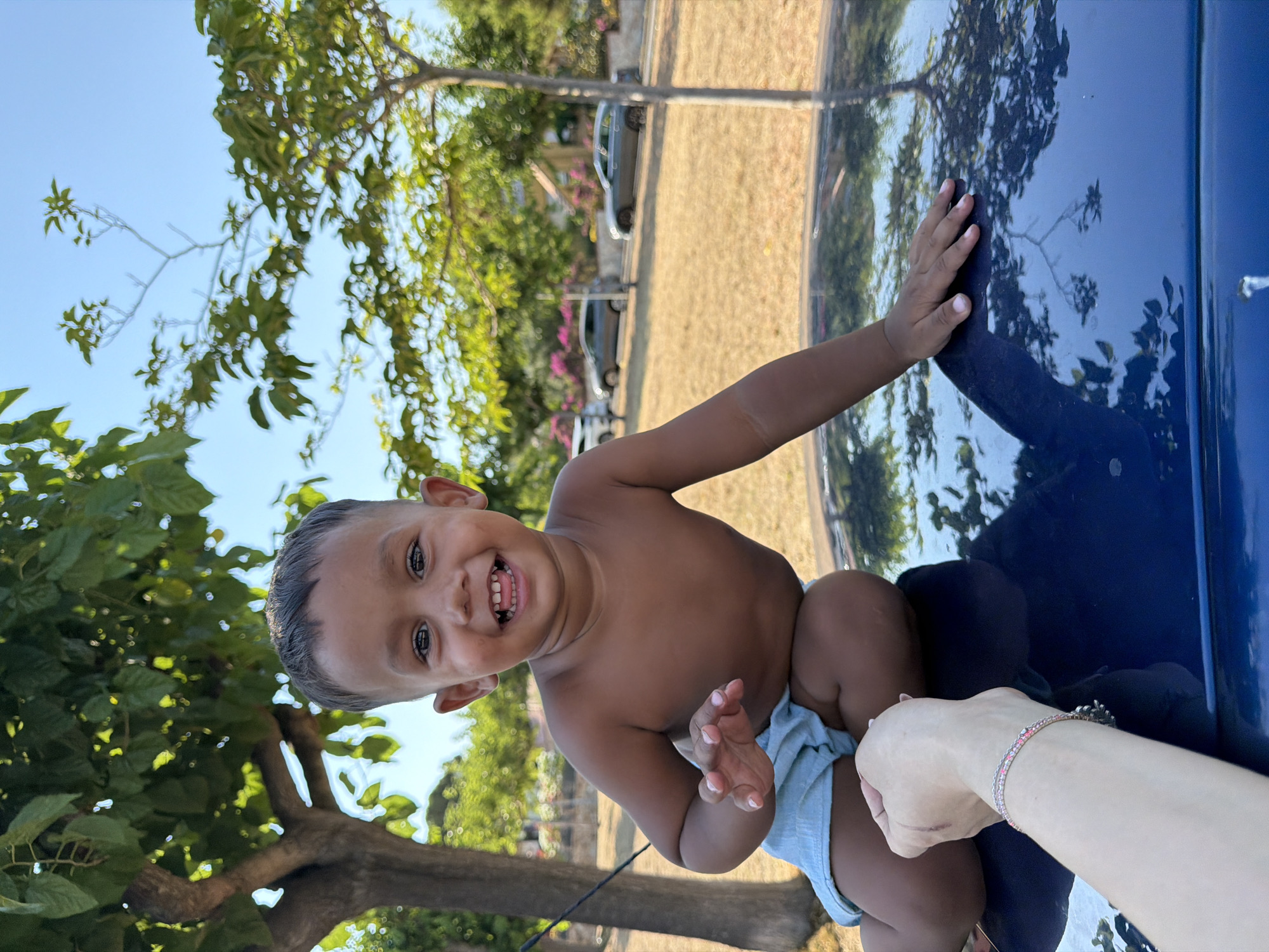 The image shows a young child, likely a toddler, sitting on the hood of a dark blue car. The child is shirtless and wearing light blue shorts. They are smiling broadly, with their mouth open and teeth visible, suggesting a moment of joy or amusement. The child's gaze is directed towards the camera, and their right hand is resting on the car, while their left hand is slightly raised.
The background of the image depicts an outdoor scene. There are trees with green leaves, and the ground appears to be dry, covered in grass and dirt. In the distance, there are some parked cars and possibly buildings. The sky is clear and blue, indicating it is daytime with fair weather. The lighting suggests it could be a sunny afternoon. The reflection on the car's surface shows a distorted image of the surroundings, including trees and the sky.
A person's hand with a bracelet is visible in the foreground, reaching towards the child. The bracelet appears to have small pink and clear stones. The overall mood of the image is cheerful and candid, capturing a happy moment with a child outdoors. The location context provided is Blanes, Spain. There is no visible text in the image.