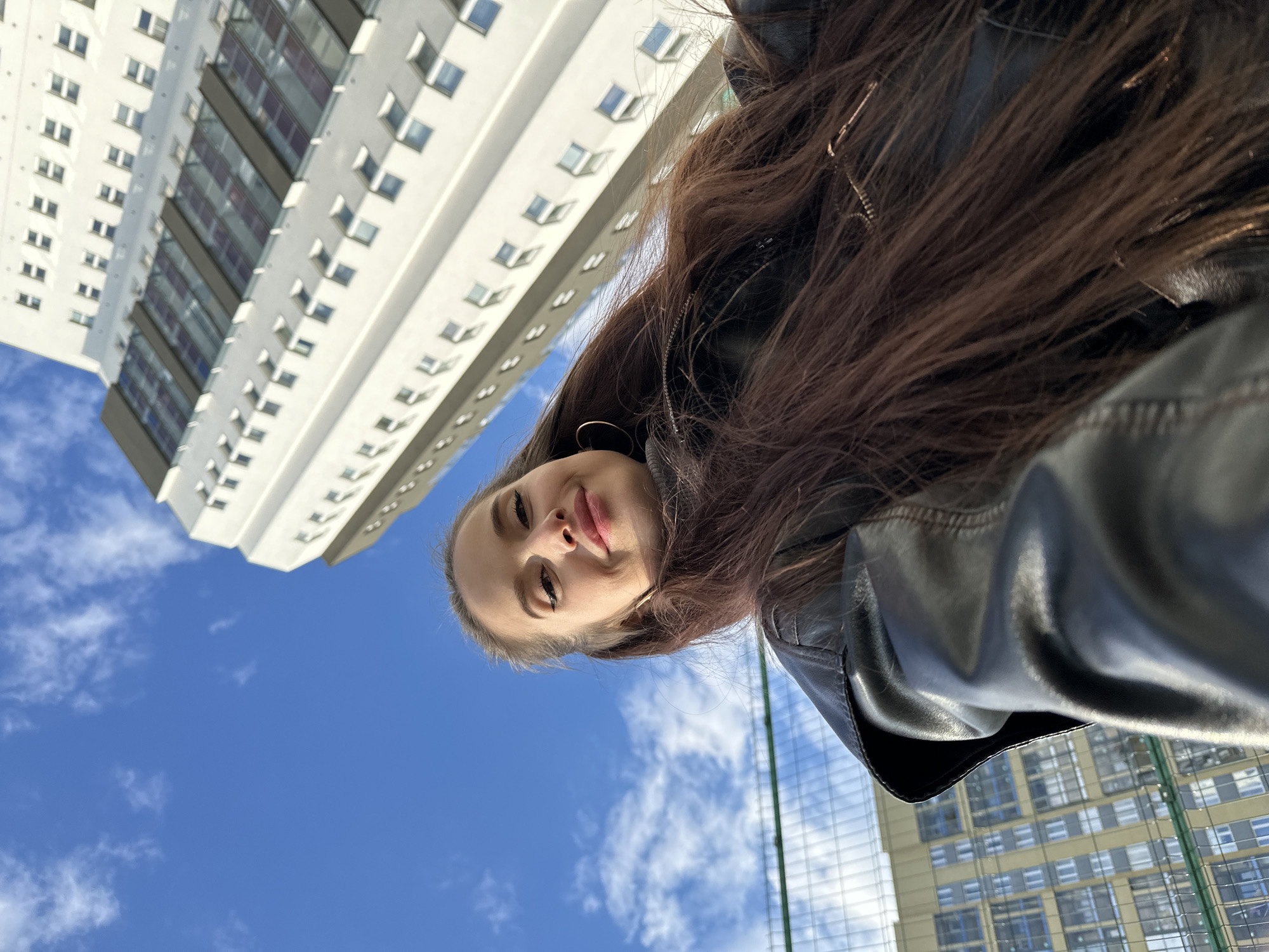 A low-angle, upward-facing shot captures a young woman looking directly at the camera, with a vibrant blue sky and tall urban buildings as her backdrop. The woman, positioned in the middle-left of the frame, has long, dark brown hair flowing downwards. She wears a black leather jacket and small, gold-colored hoop earrings. Her expression is calm with a slight smile, and her lips are subtly tinted.
The photo's unique perspective emphasizes the towering structures around her. To the left and behind her, a large, predominantly white building with numerous small, rectangular windows dominates the upper-left quadrant. Some windows appear slightly open. Another building, with a lighter beige or yellowish hue and a grid-like pattern of windows, is visible in the lower-right background, partially obscured by a green mesh fence or netting. This netting extends from the lower-right towards the center-right.
The scene is set outdoors on a bright day, likely midday or early afternoon, under a clear blue sky dotted with scattered white clouds. The strong sunlight illuminates the buildings and the woman's face. The urban environment, characterized by modern high-rise architecture and the bright, clear weather, suggests a vibrant city setting in Yekaterinburg, Russia. There is no visible text in the image.