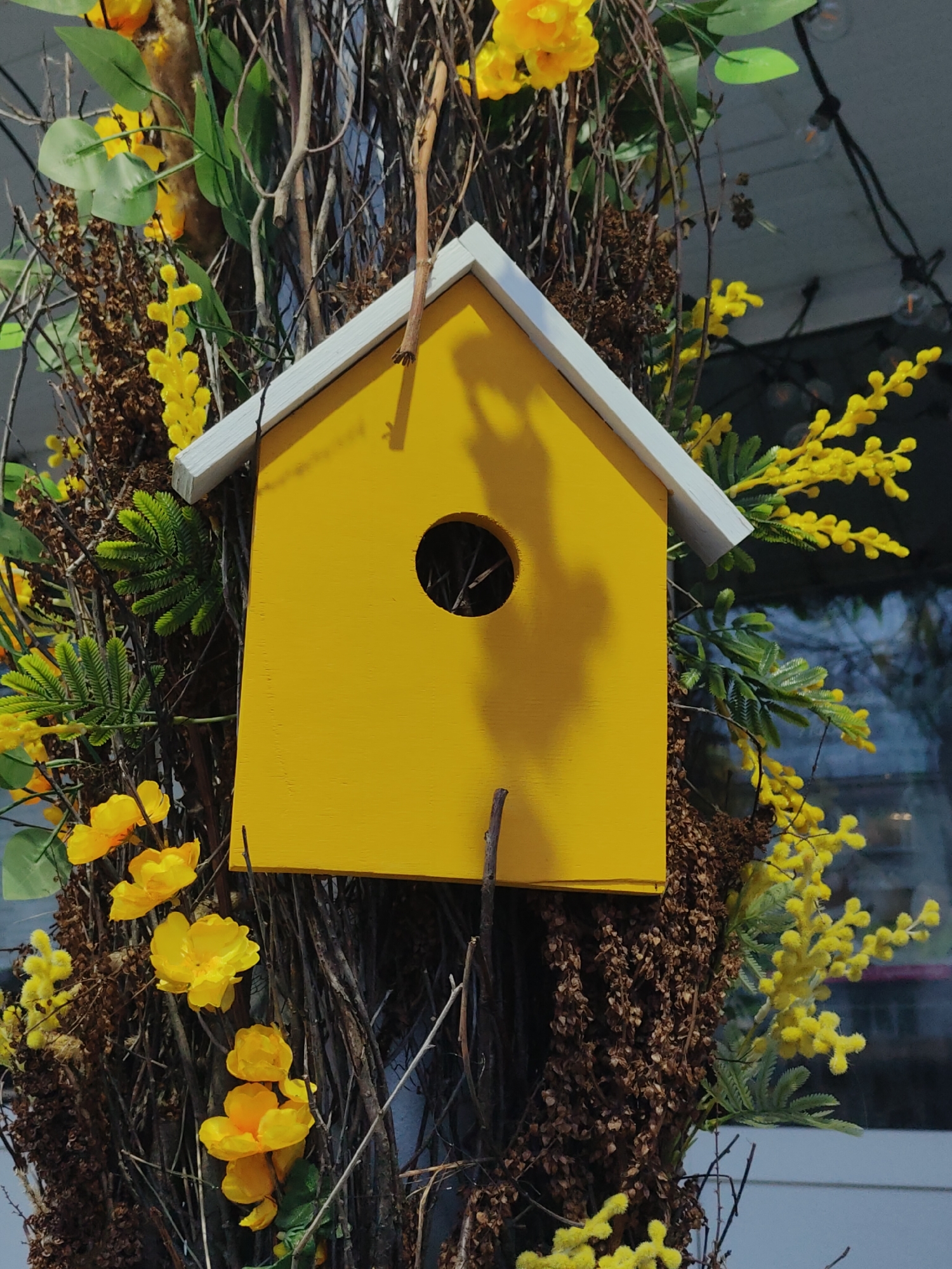 A medium shot depicts a yellow birdhouse with a white roof, surrounded by branches and yellow flowers. The birdhouse has a circular opening. The scene is likely in Chaikovsky, Russia, based on implied geographic context.