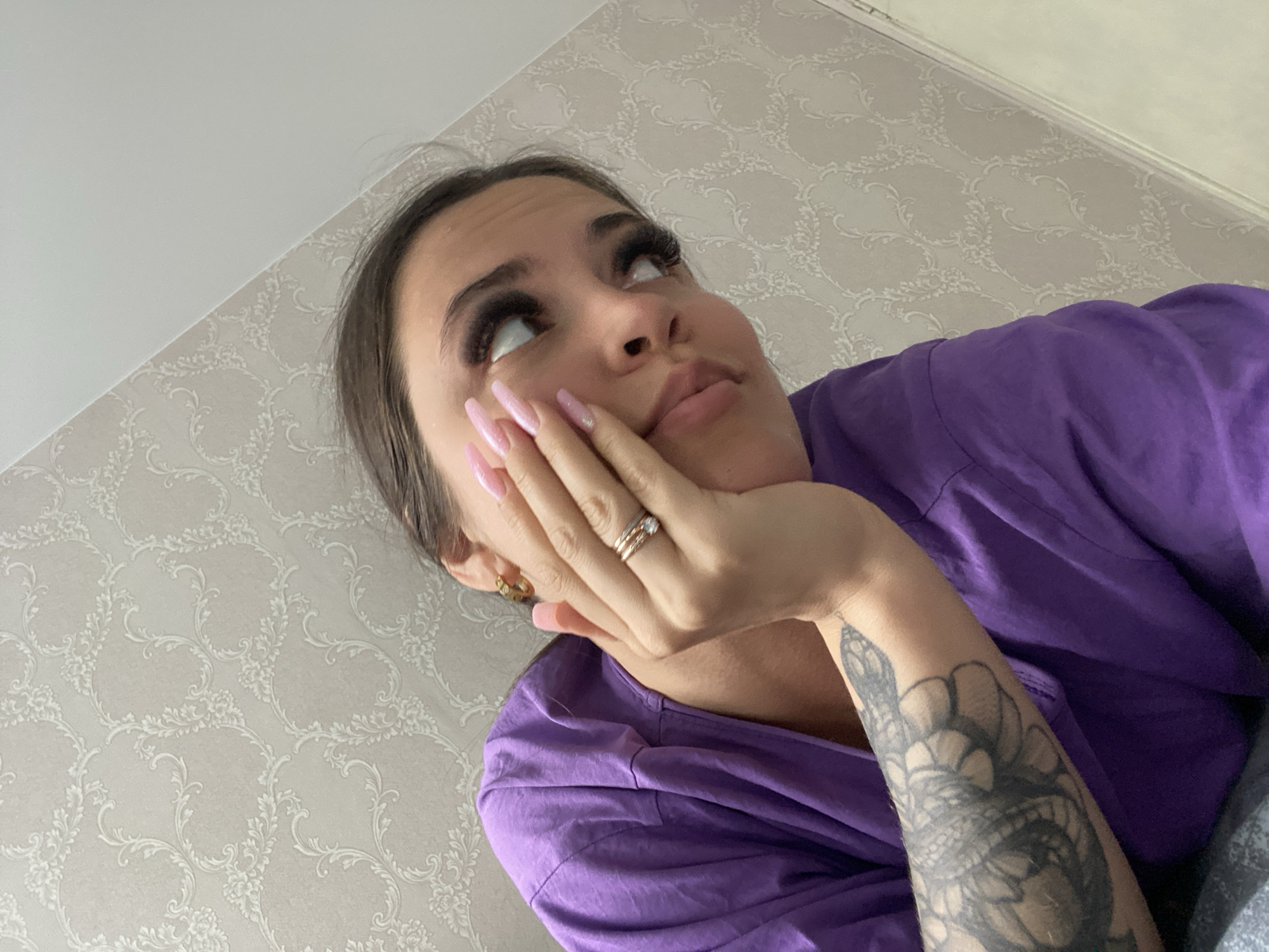 A young woman is captured in a close-up, low-angle shot, looking upwards and to the right with a pensive or thoughtful expression, her left hand supporting her chin. She has long, dark hair, prominent long false eyelashes, and well-manicured, long, light pink fingernails. On her left ring finger, she wears a ring composed of several intertwined bands. A small gold hoop earring is visible in her left ear. She is wearing a purple, possibly short-sleeved, top. A distinct tattoo, featuring a snake intertwined with what appears to be floral or scale-like patterns, is visible on her right forearm. The background consists of a wall covered in a light grey or beige wallpaper with an intricate, repeating pattern of white or cream floral and scroll motifs. Above the patterned wallpaper, a plain off-white ceiling or upper wall section is visible. The overall setting is indoors, likely a private room in Chaikovsky, Russia. No visible text is present, and the time of day is not discernible.