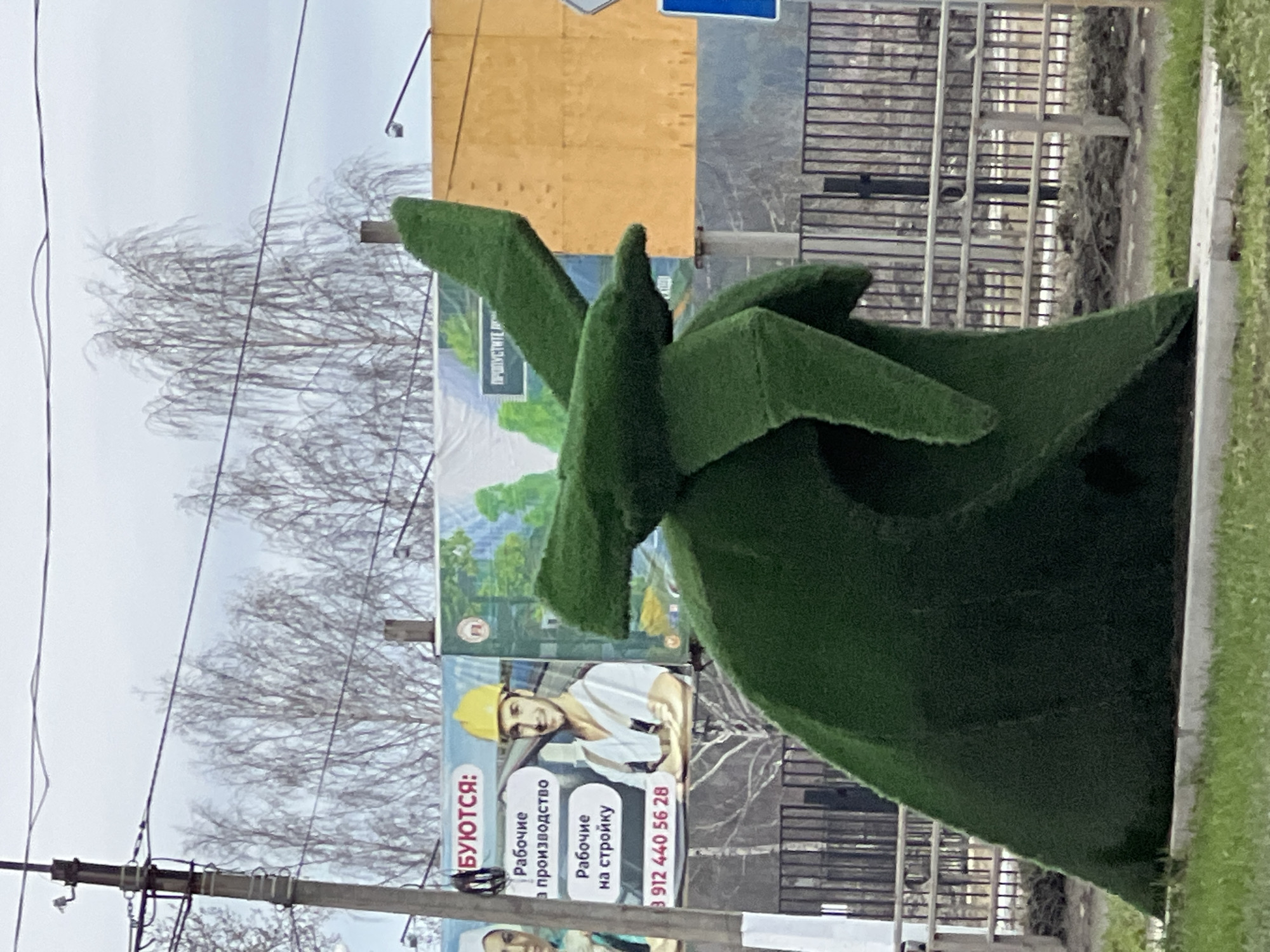 The image captures a roadside scene in Chaikovsky, Russia. A large, abstract sculpture covered in green artificial turf dominates the foreground, likely a public art installation. Behind the sculpture is a series of billboards advertising construction workers, with Russian text and phone numbers. Bare trees and power lines are visible against an overcast sky. A section of fencing runs behind the billboards. The sculpture is positioned on a concrete curb.