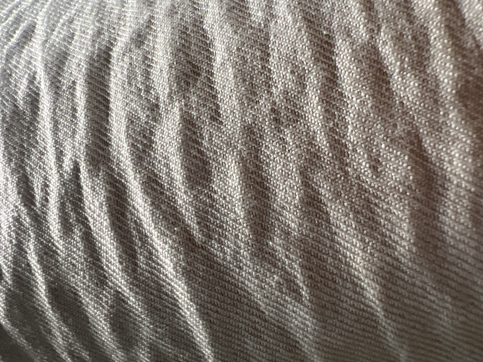 The image presents a high-resolution, close-up photograph of a textile surface, devoid of any discernible objects other than the fabric itself. The material exhibits a fine, diagonal twill weave pattern, with individual threads appearing as small, interlocking components. The fabric's color is a muted greige or light brownish-grey. Strong, directional lighting illuminates the scene from the upper left, casting distinct, elongated shadows that emphasize the three-dimensional texture of the weave and the multiple gentle, horizontal undulations or folds present across the fabric. These folds create alternating bands of bright highlights and deeper shadows, accentuating the textile's topographic variations. No people, actions, or specific environmental features are visible to denote a particular location within the city of Casablanca, Morocco.