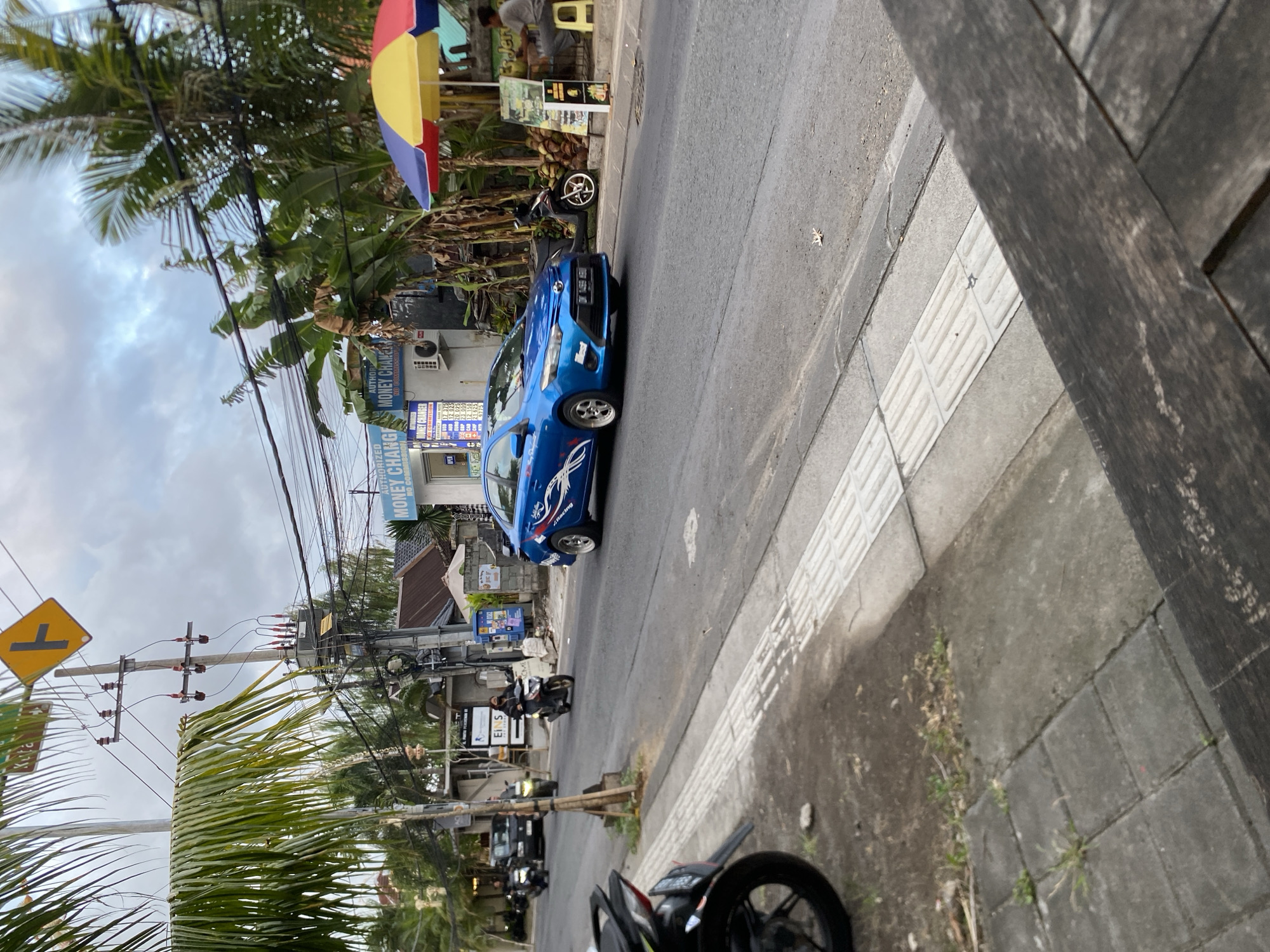 The image depicts a street scene in Mengwi, Indonesia, likely during daylight hours under an overcast sky. A blue sedan with white racing stripes is driving on the right side of the road. In the background, several motorcycles are visible, with two riders on one, one on another, and one rider on a third. Buildings line the street, including one with "MONEY CHANGER" signage. Palm trees and other foliage are abundant, suggesting a tropical environment. A person is seated under a colorful umbrella near a pile of coconuts. The foreground shows a sidewalk and a road with a white line marking.
