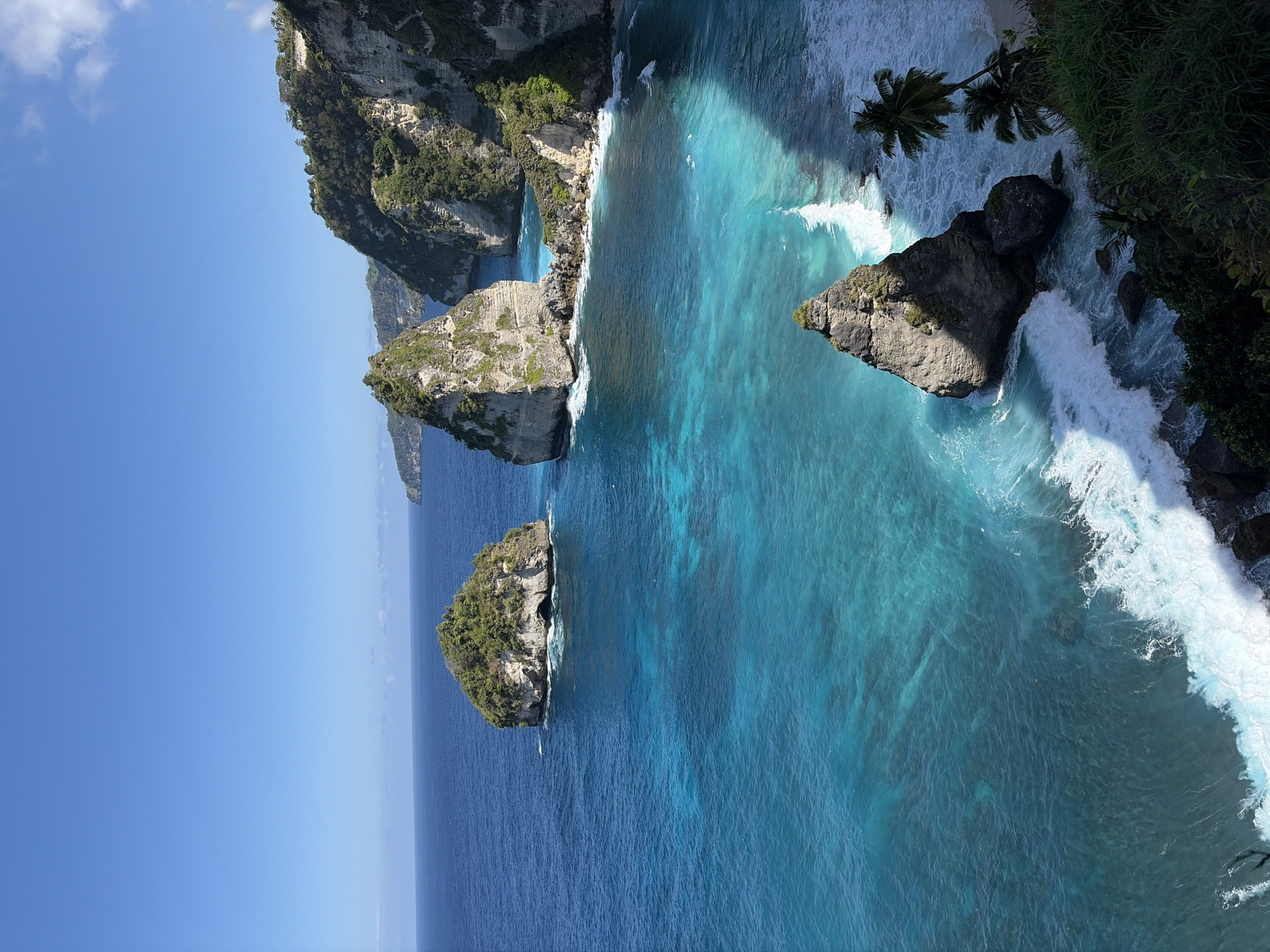 This is a high-angle, wide-shot, eye-level photograph of the coastline at Diamond Beach in Nusa Penida, Indonesia. The scene is dominated by the vivid turquoise waters of the ocean meeting rugged, rocky cliffs. Several large rock formations jut out of the sea, some covered in lush green vegetation. Waves crash against the rocks and the sandy beach, creating white foam. The sky is a clear, bright blue with a few wispy clouds. A small section of a sandy beach is visible in the lower right corner, along with palm trees and dense green foliage. There are no people visible in the image. The sun casts strong shadows across the water and the rock formations. The overall impression is one of a dramatic and beautiful coastal landscape.
