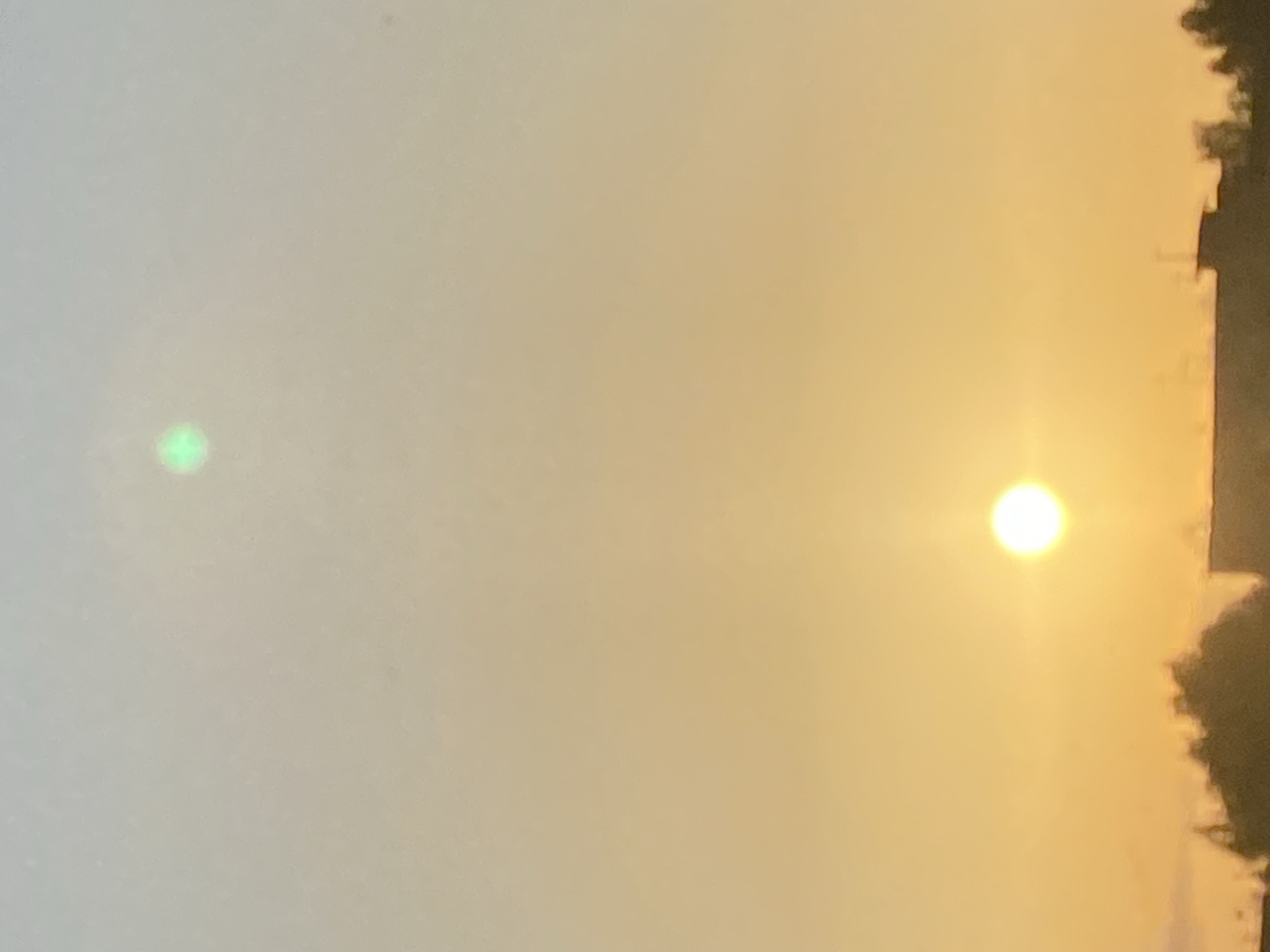The image displays a dusky sky with the sun low on the horizon, casting a warm, orange glow. The lower portion of the image shows the silhouettes of buildings and trees. In the sky, two bright, round objects are visible. The lower object is a clear, bright white circle with a distinct halo, representing the sun. Above and slightly to the left of the sun, a smaller, greenish, circular object is present, which appears to be a lens flare or another optical artifact. The sky transitions from a pale blue at the top to a rich orange near the horizon.