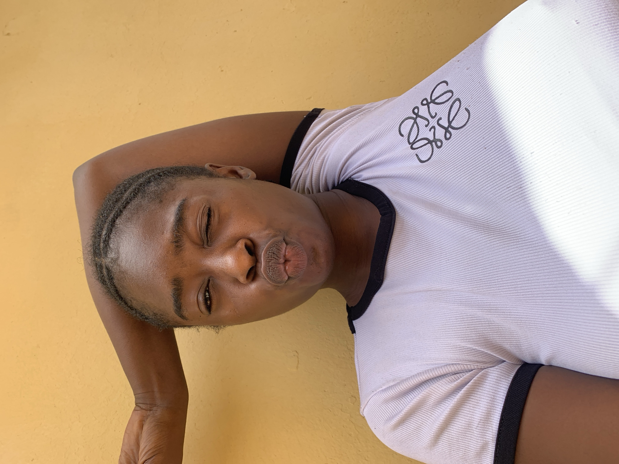 A person with dark skin and braided hair is posed against a yellow wall. The person is wearing a white t-shirt with black trim and a black logo. They are winking with one eye closed and making a kissing face with their lips. The photo is taken in Rafin Par, Nigeria, but specific location details are not discernible.
