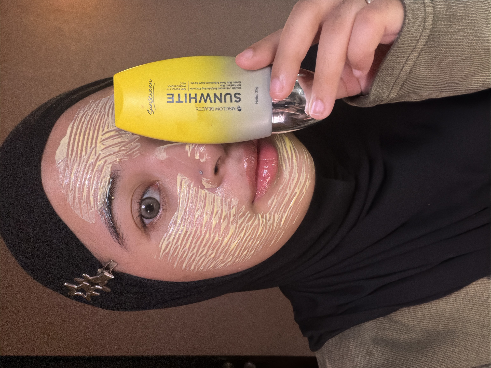 A close-up, tilted shot captures a person, likely in East Jakarta, Indonesia, in the midst of applying a cosmetic product to their face. The subject is wearing a black hijab and an olive green top, visible on the right side of the frame. Their face is partially covered with streaks of a yellowish product, indicating an ongoing application process. One eye, looking directly at the viewer, and glossy lips are visible, along with a nose piercing.
The person holds a yellow and gray tube of "MS GLOW BEAUTY SUNWHITE" sunscreen to their face. The visible text on the product includes "Sunscreen," "MSGLOW BEAUTY," "SUNWHITE," "Double Advanced Brightening Formula for Radiant Skin," "Evens Skin Tone & Reduces Dark Spots," "SPF 50PA++++ WhiteCellDNA," and "Vit C," with a net weight of "Netto: 35g."
A silver-colored, star-shaped brooch or accessory is pinned to the hijab near the person's left temple. The background appears to be an indistinct, dark reddish-brown surface, suggesting an indoor setting. The image is taken at an angle, with the person's head rotated counter-clockwise.